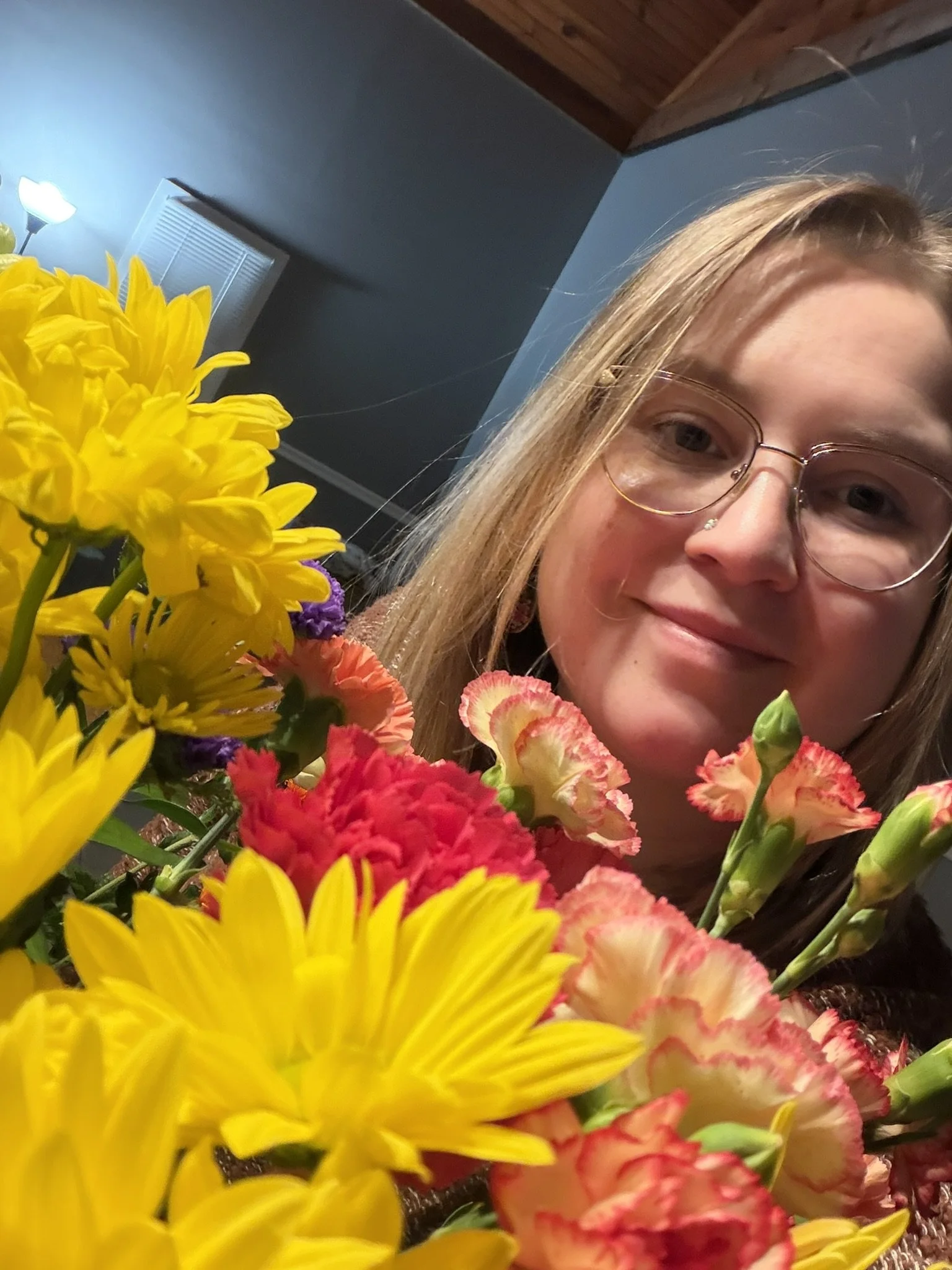 A young woman with glasses and long blonde hair is smiling behind a colorful bouquet of yellow, pink, and purple flowers in an indoor setting.
