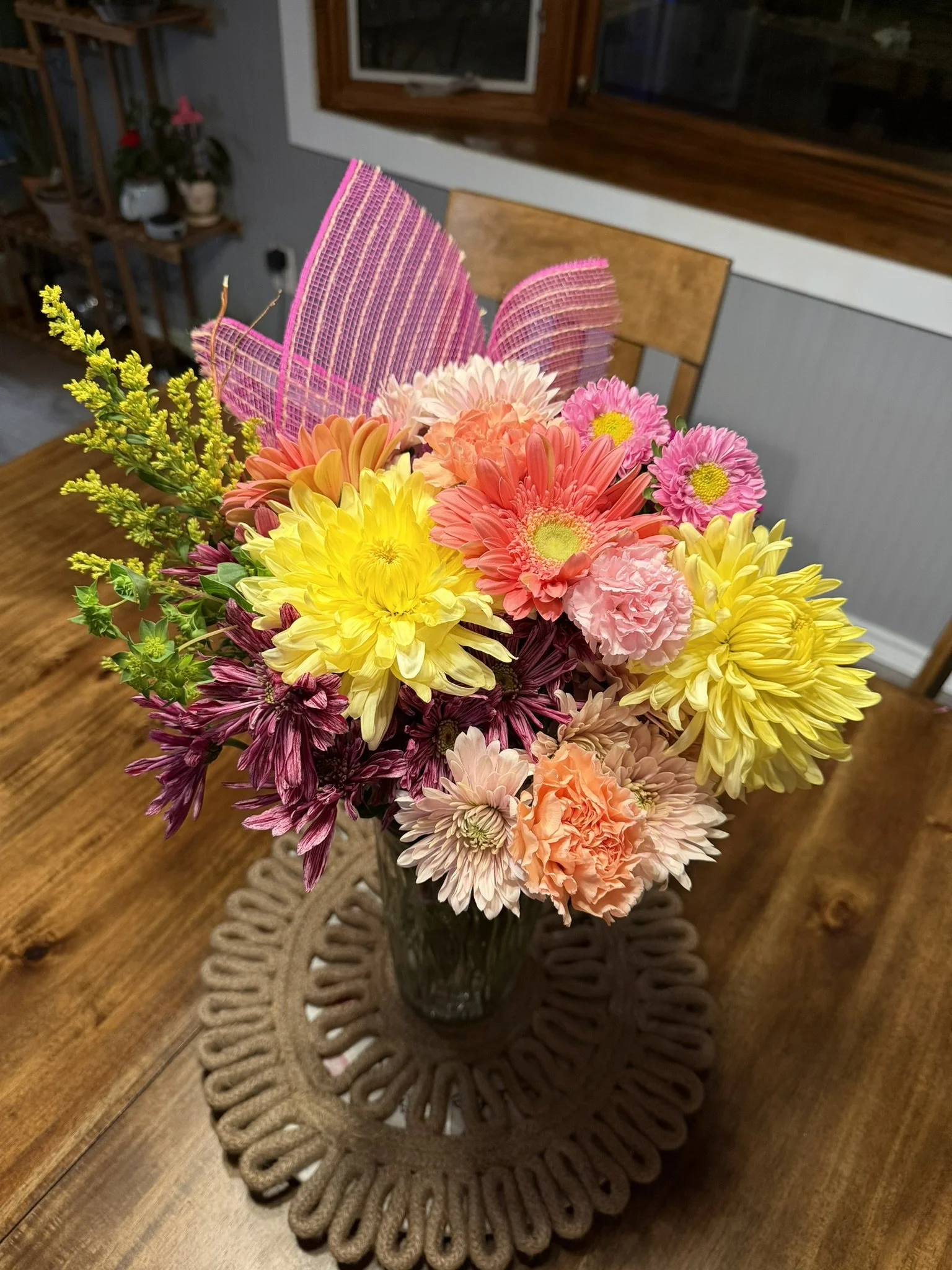 A colorful bouquet of flowers on a wooden table, with pink, yellow, and peach blooms, including daisies and carnations, arranged in a glass vase, with a decorative placemat underneath.