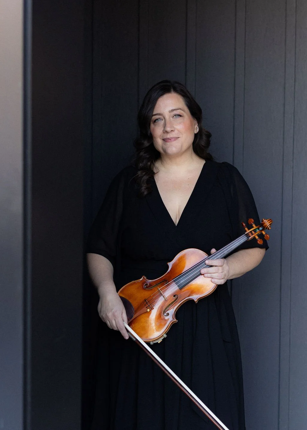 A woman with dark hair in a black dress holding a violin and bow, standing against a dark wall.