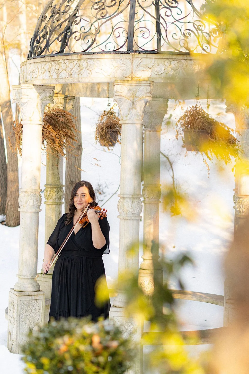 A woman in a black dress playing a violin outdoors near a decorative gazebo with hanging planters in a snow-covered setting.