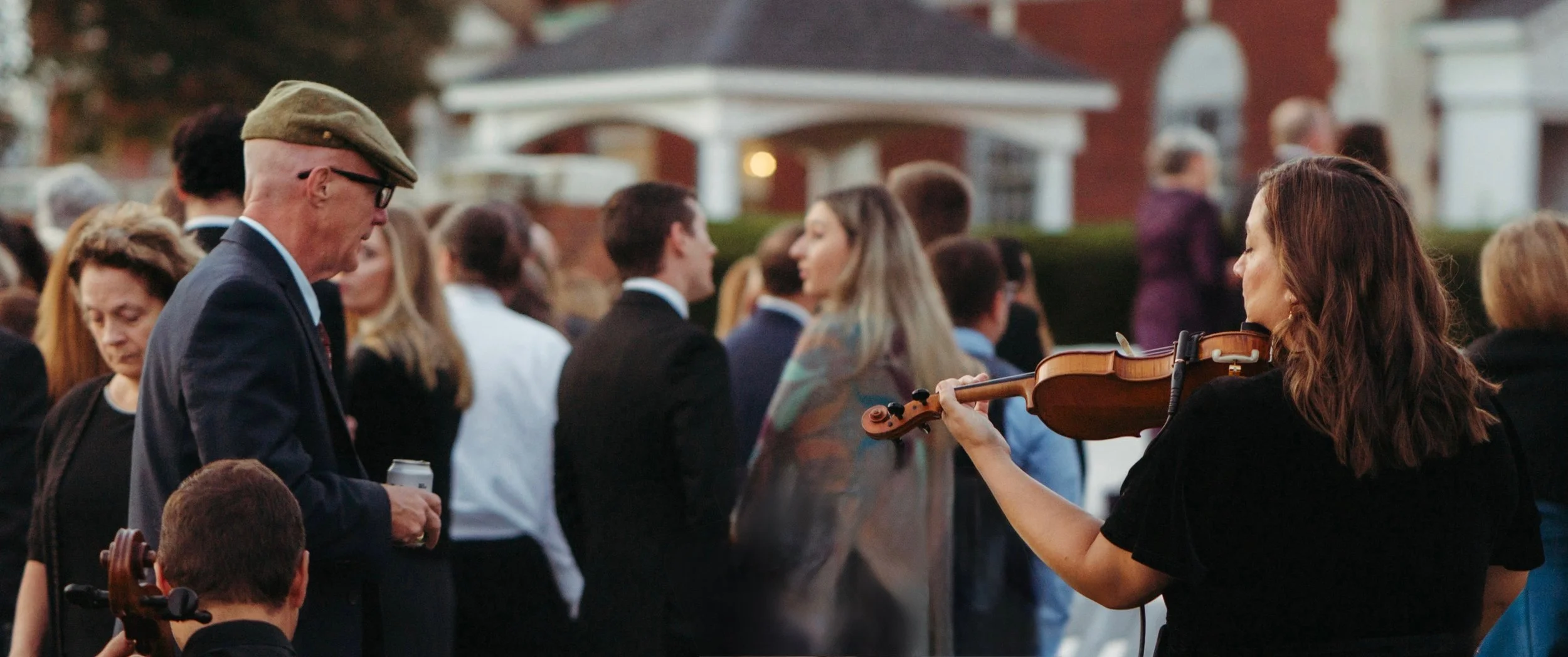 A woman playing the violin at an outdoor gathering with several people in formal attire socializing in the background.