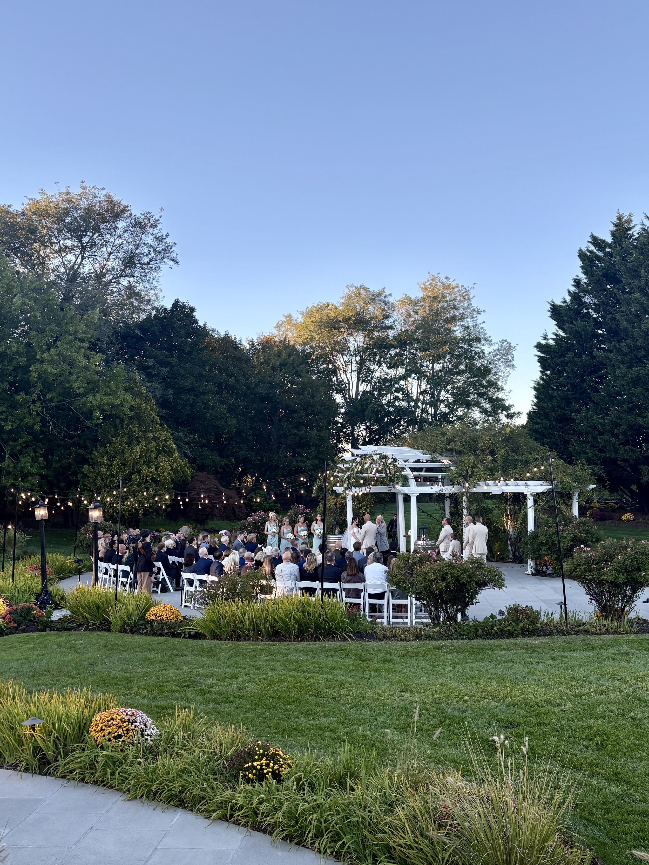 An outdoor wedding ceremony taking place on a green lawn during the evening, with guests seated in white chairs facing a white wedding arch. String lights are hanging above, and there are trees and shrubs around the area.
