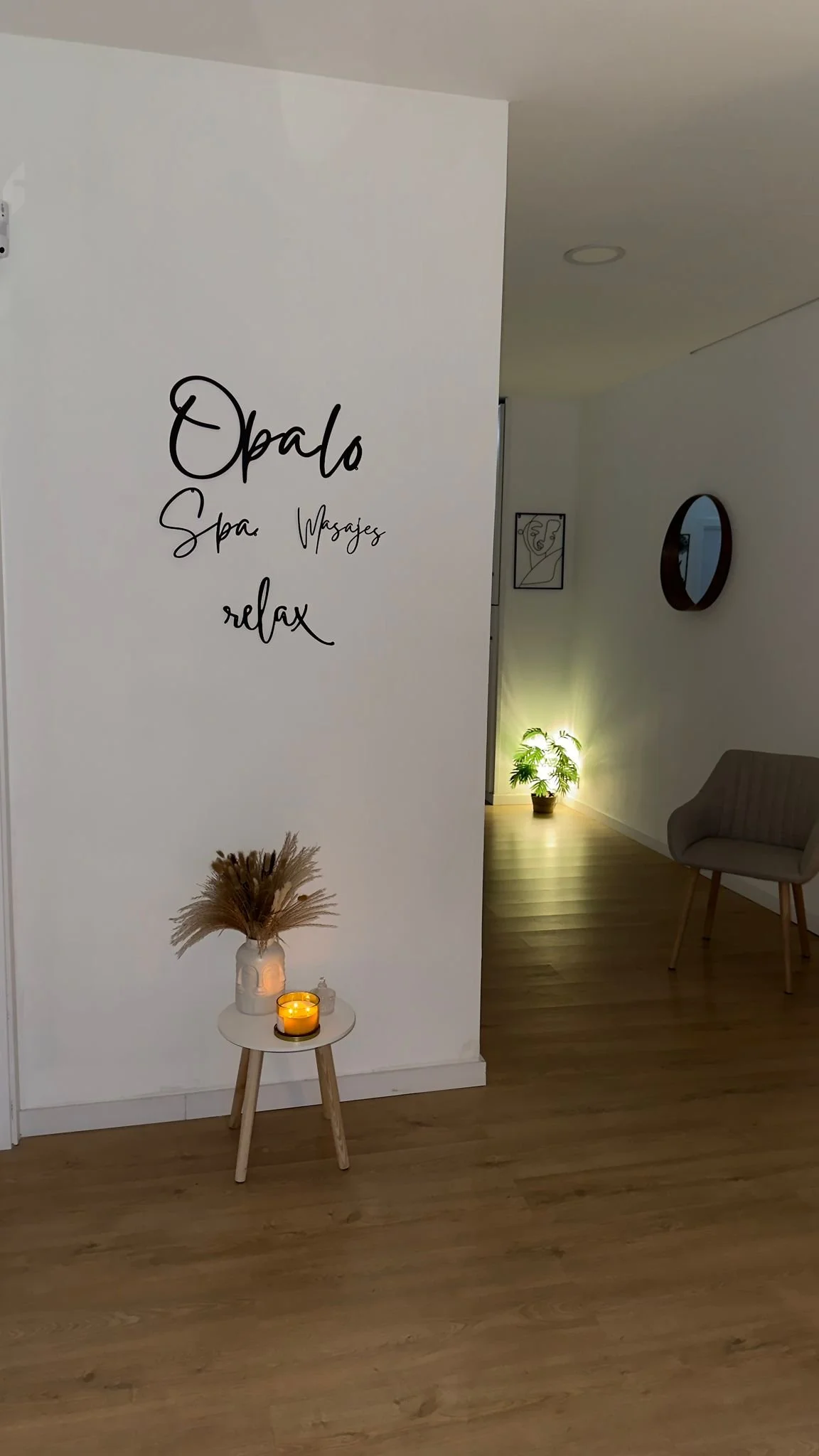 Interior de un spa con pared blanca y letrero que dice "Opalo Spa Wwagyaz relax", una mesa pequeña con una vela y decoración de paja seca, y en el fondo plantas y cuadros decorativos en la pared.