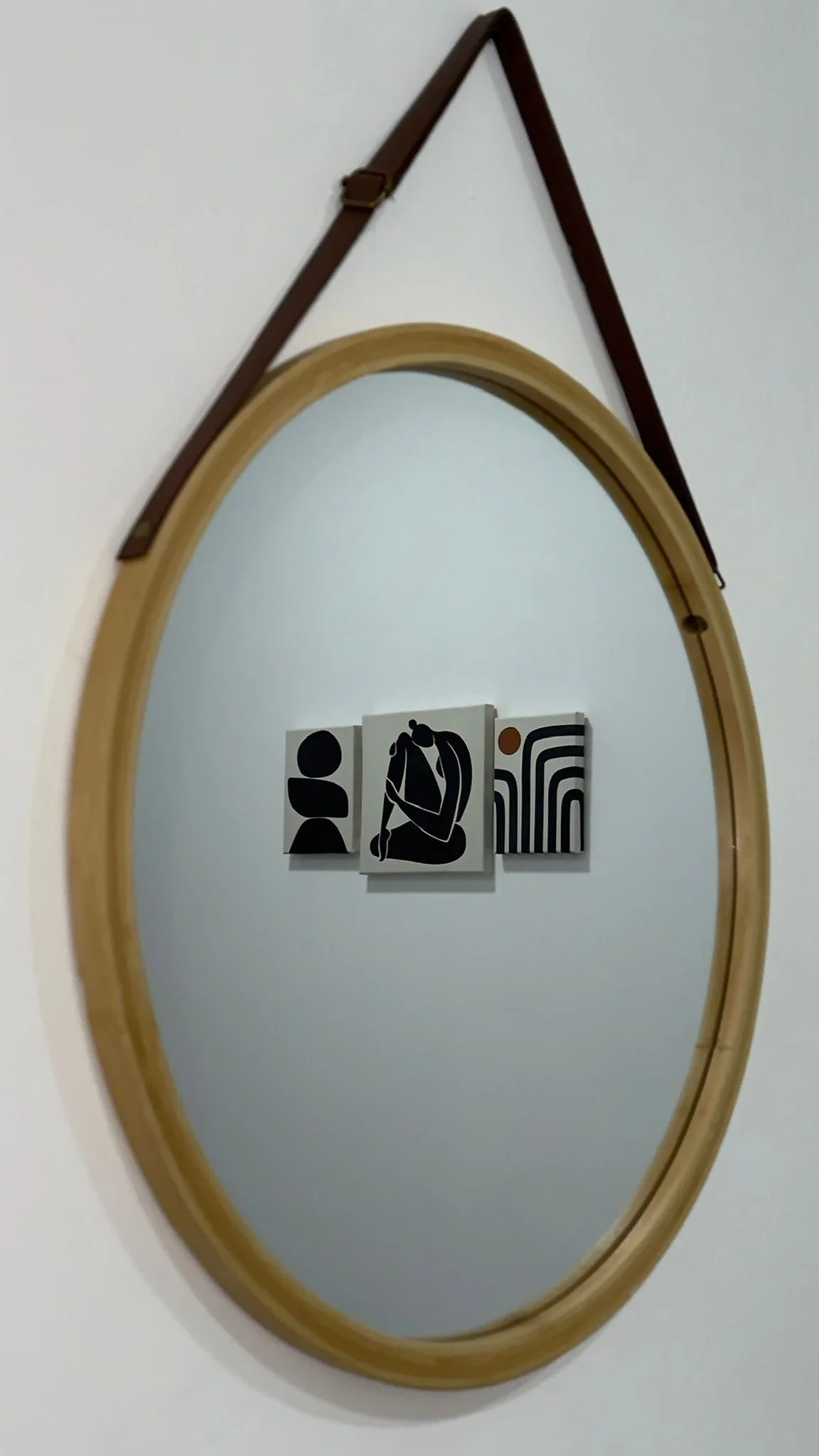 Espejo de marco ovalado con cinta de cuero en la parte superior, colgado en la pared, reflejando tres cuadros abstractos en la pared.