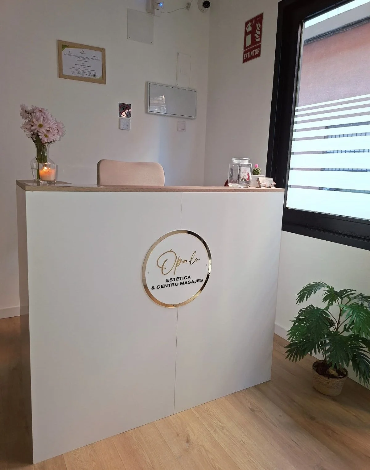 Mostrador de recepción en un centro de estética llamado 'Opalo', decorado con flores y una vela, y con un cartel de 'No fumar' en la pared detrás, junto a un certificado enmarcado y un signo de incendio.