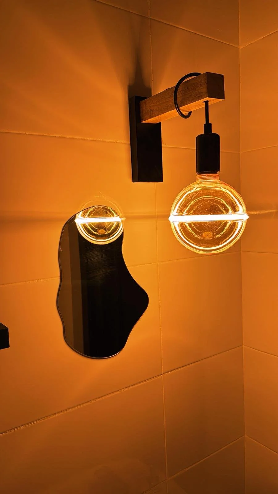 Lámpara de pared con bombilla de estilo industrial. Reflejo de la bombilla en un espejo pequeño en la pared. Fondo de azulejos beige.