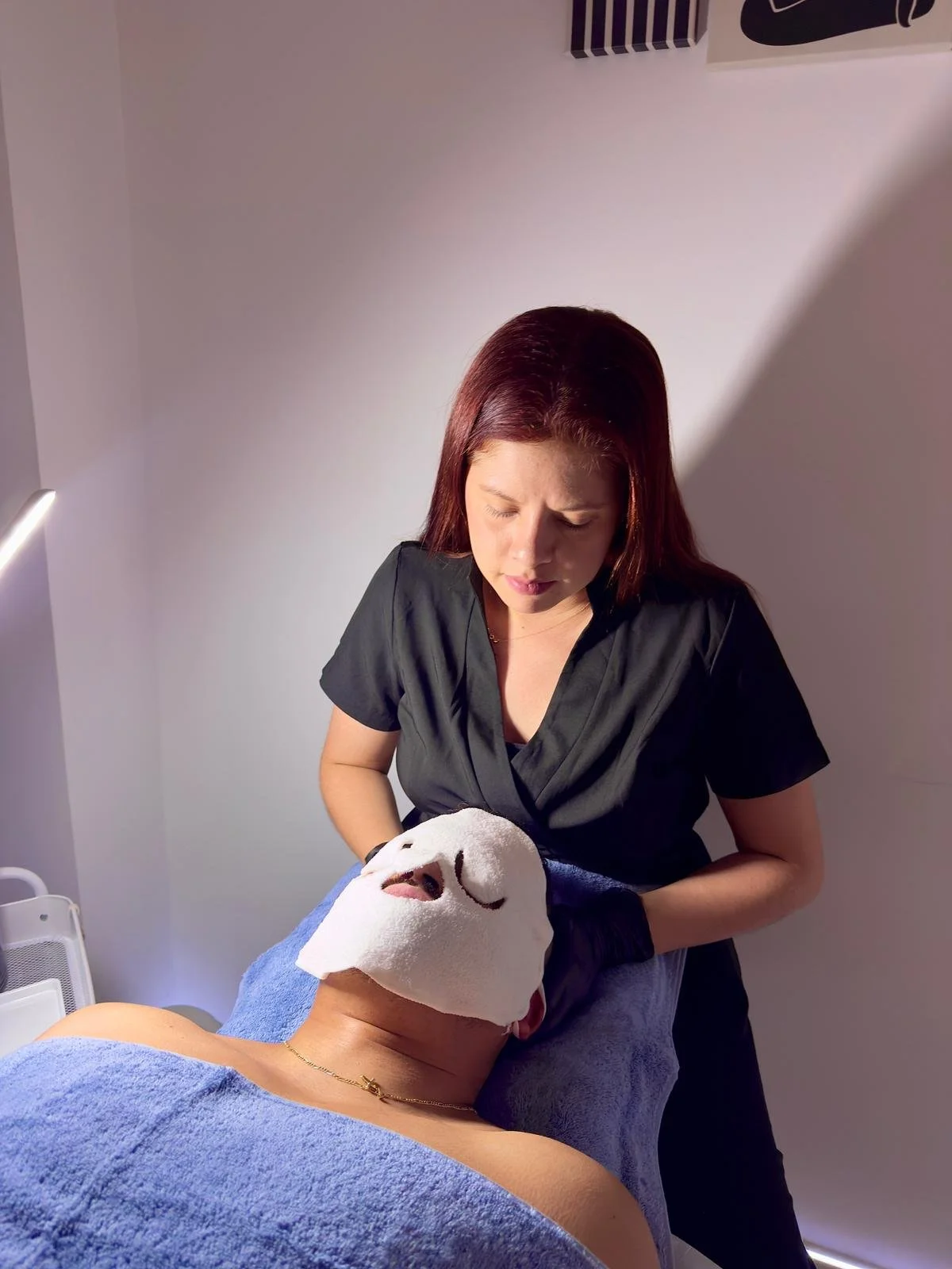 Una mujer atendiendo a otra en una cama en una clínica de estética o spa, con una máscara facial y una toalla azul en el cuerpo.