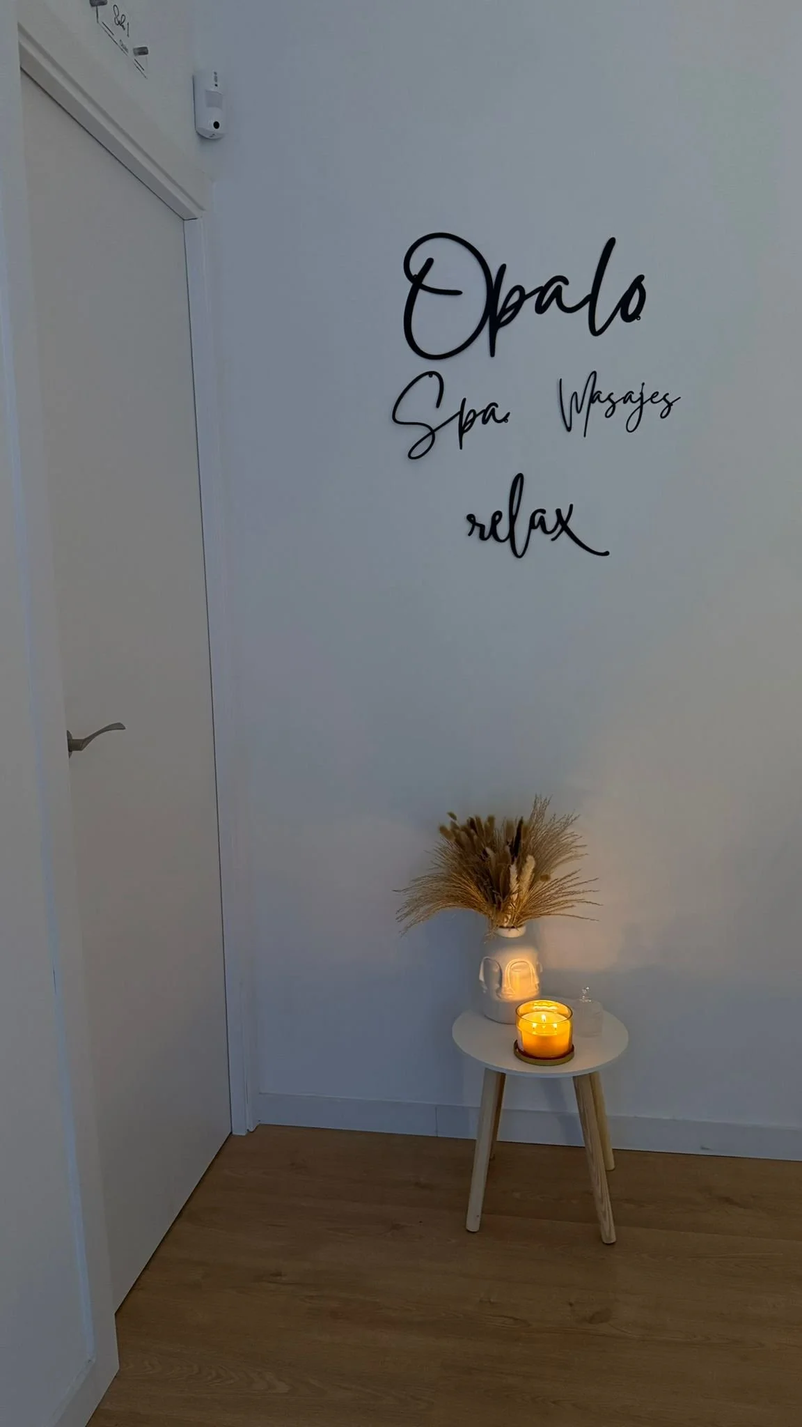 Espacio con pared blanca, cartel que dice 'Opalo Spa. Masajes. relax' en letras negras, mesa pequeña con vela encendida y jarrón con flores secas.