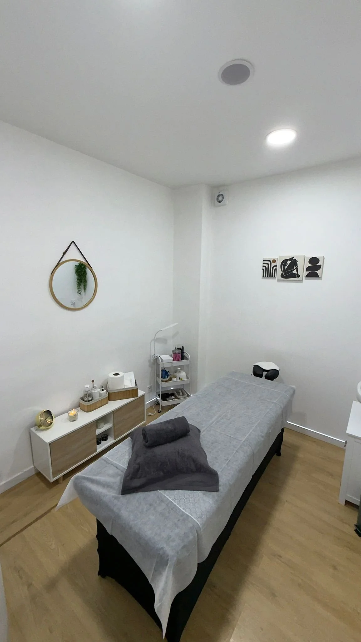 Sala de masaje con camilla cubierta con sábanas blancas y toallas grises, decorada con arte abstracto en la pared y espejos decorativos, con muebles minimalistas y accesorios de spa.