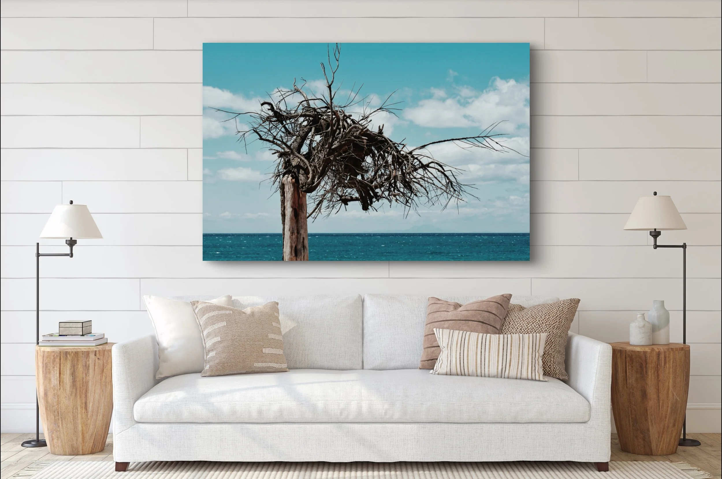 A large wall art of a leafless tree with twisted branches standing in front of a blue ocean and sky with clouds, hanging above a white couch in a modern living room.
