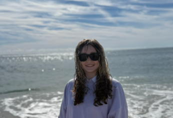Woman with curly hair and sunglasses standing on the beach with the ocean and cloudy sky behind her.
