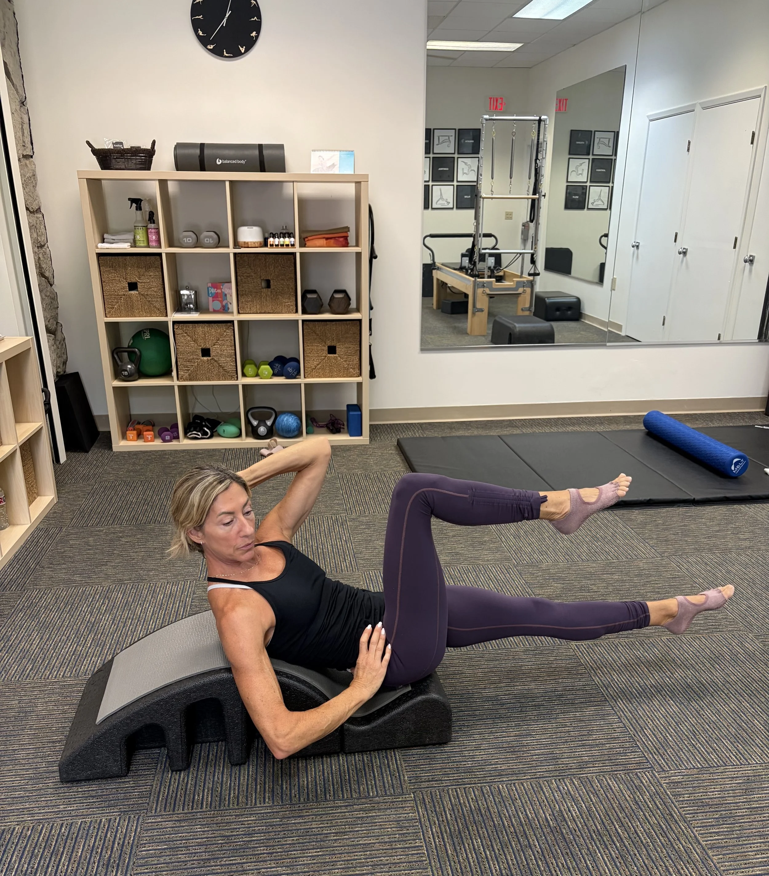 Woman lying on a black cushioned exercise step, performing a pilates exercise with her arms raised behind her head and one leg elevated, wearing purple leggings and a black tank top in a fitness room with various workout equipment and a large mirror.