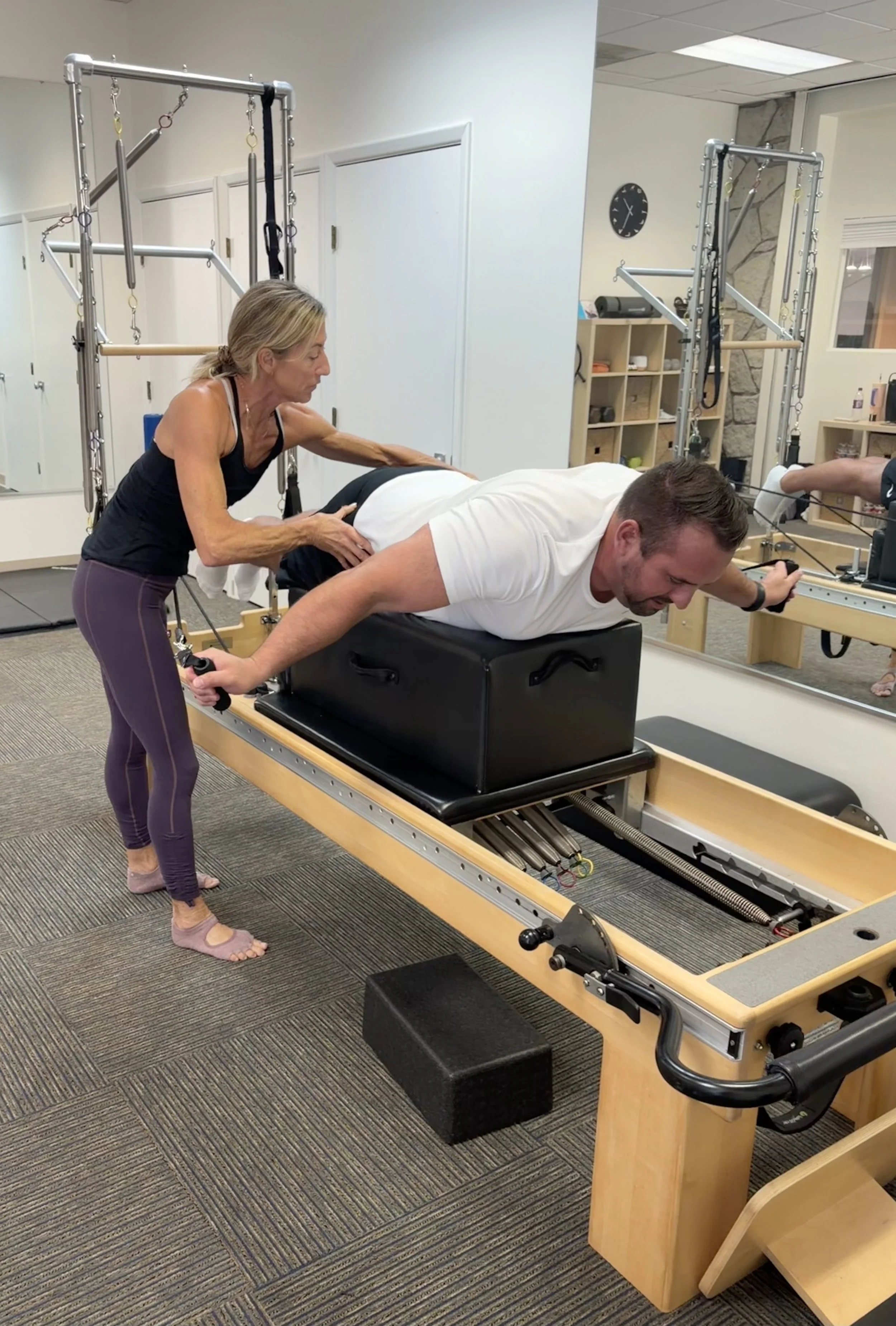 An elderly woman assisting a man in a white shirt with a physical therapy exercise on a specialized medical machine in a therapy clinic.
