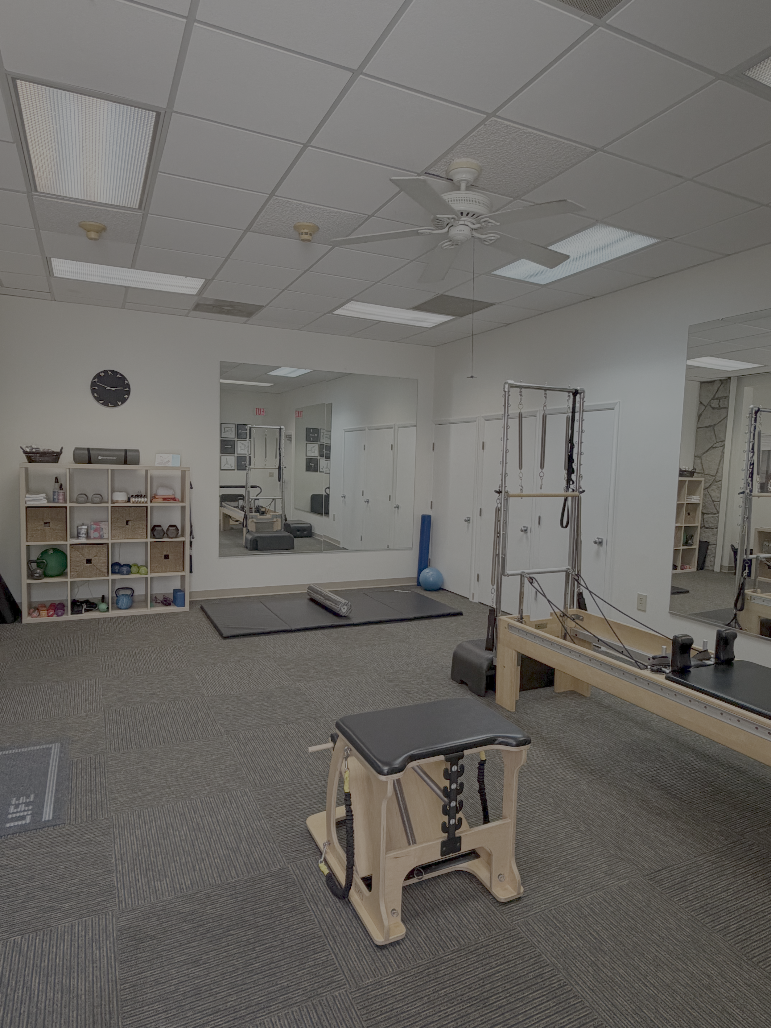 A physical therapy or fitness room with Pilates reformer and other exercise equipment, a mirrored wall, and storage shelves with various fitness accessories.