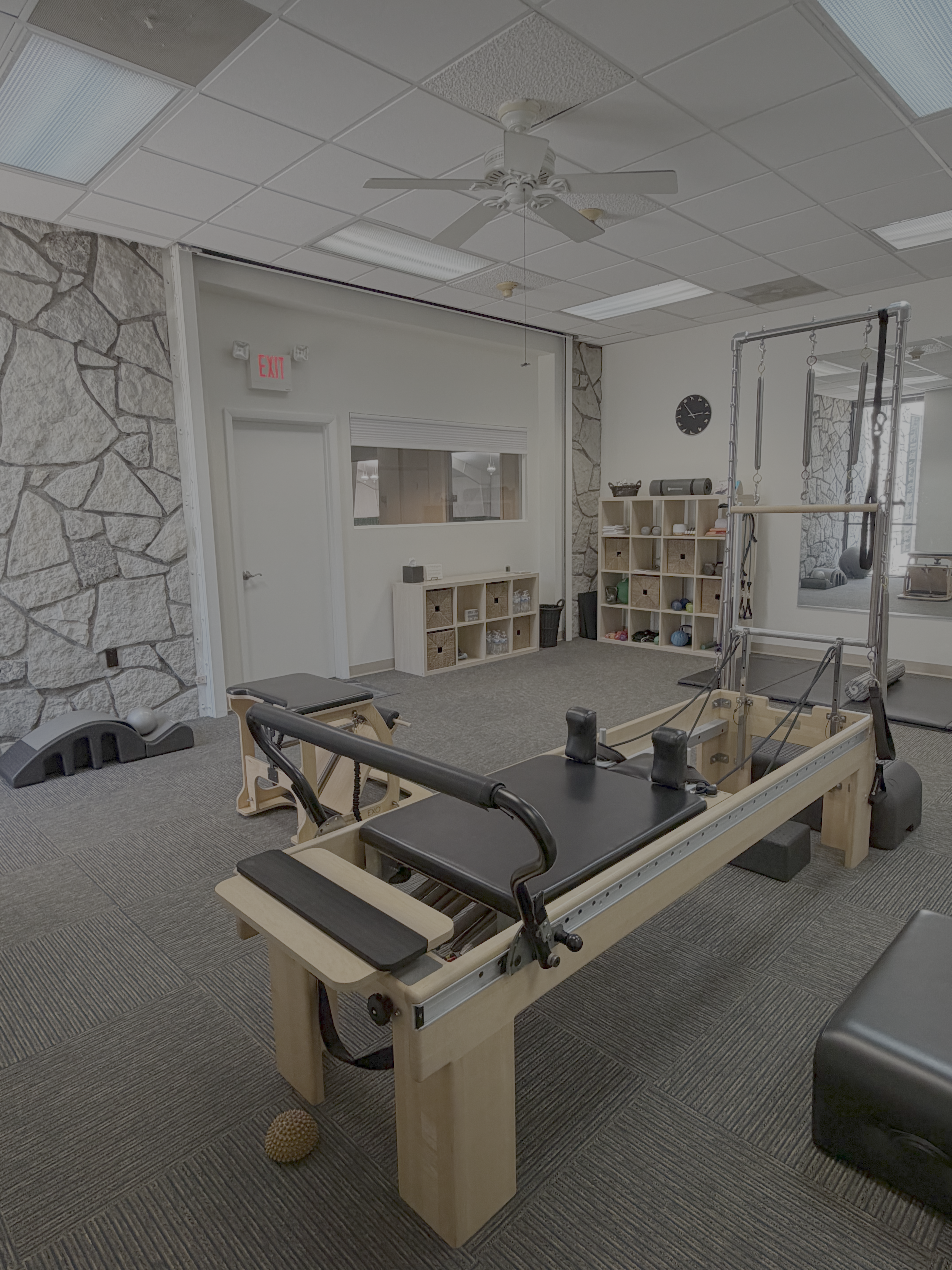 A physical therapy or rehabilitation room with a treatment table, exercise equipment, a wall clock, shelves with rounded bins and fitness gear, and a large mirror. The ceiling is equipped with fluorescent lights and a ceiling fan.