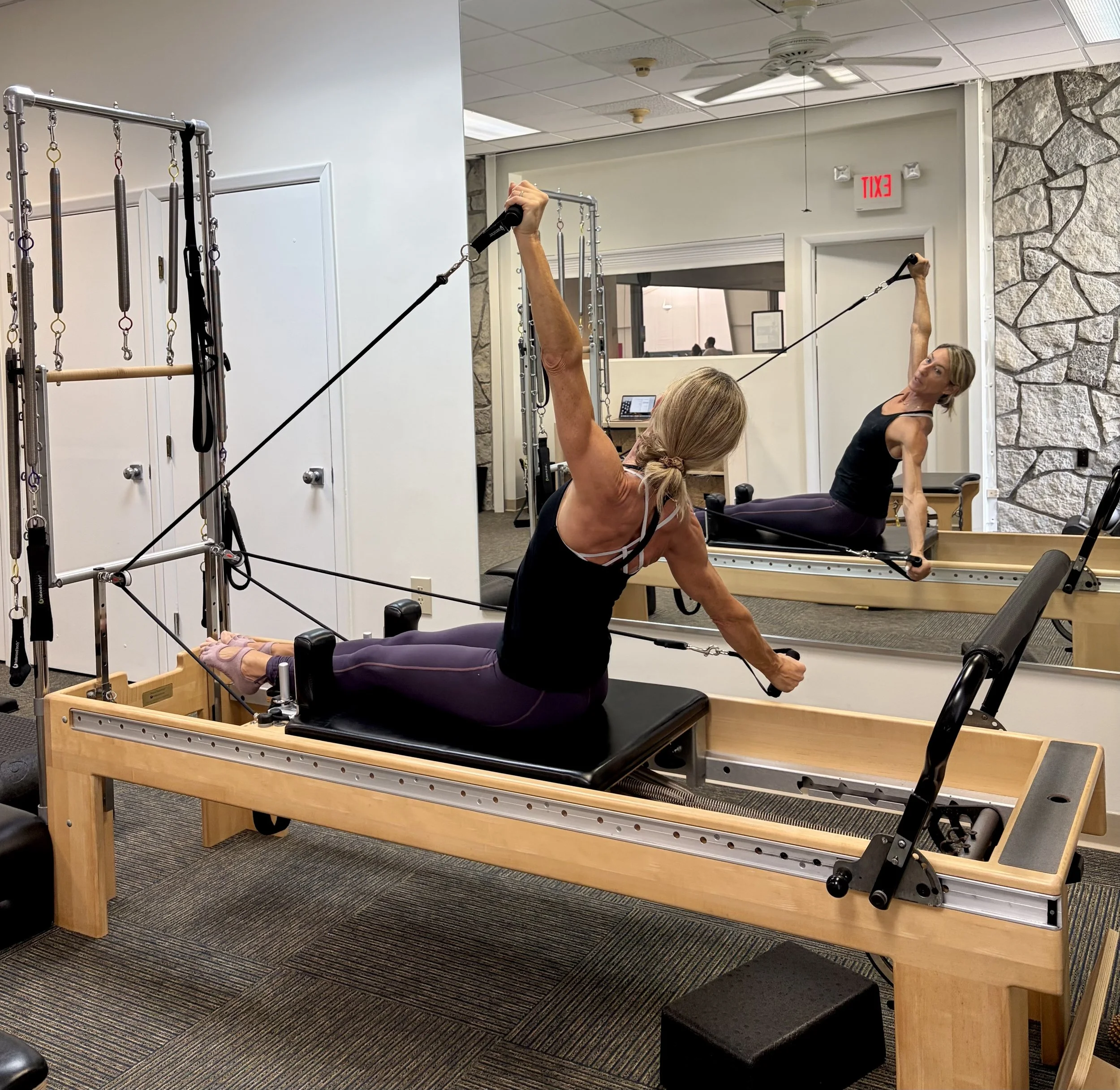 Two women working out with resistance bands and Pilates equipment in a gym studio with stone accent walls and white ceiling, one woman engaging in an arm exercise and the other woman performing a side plank.