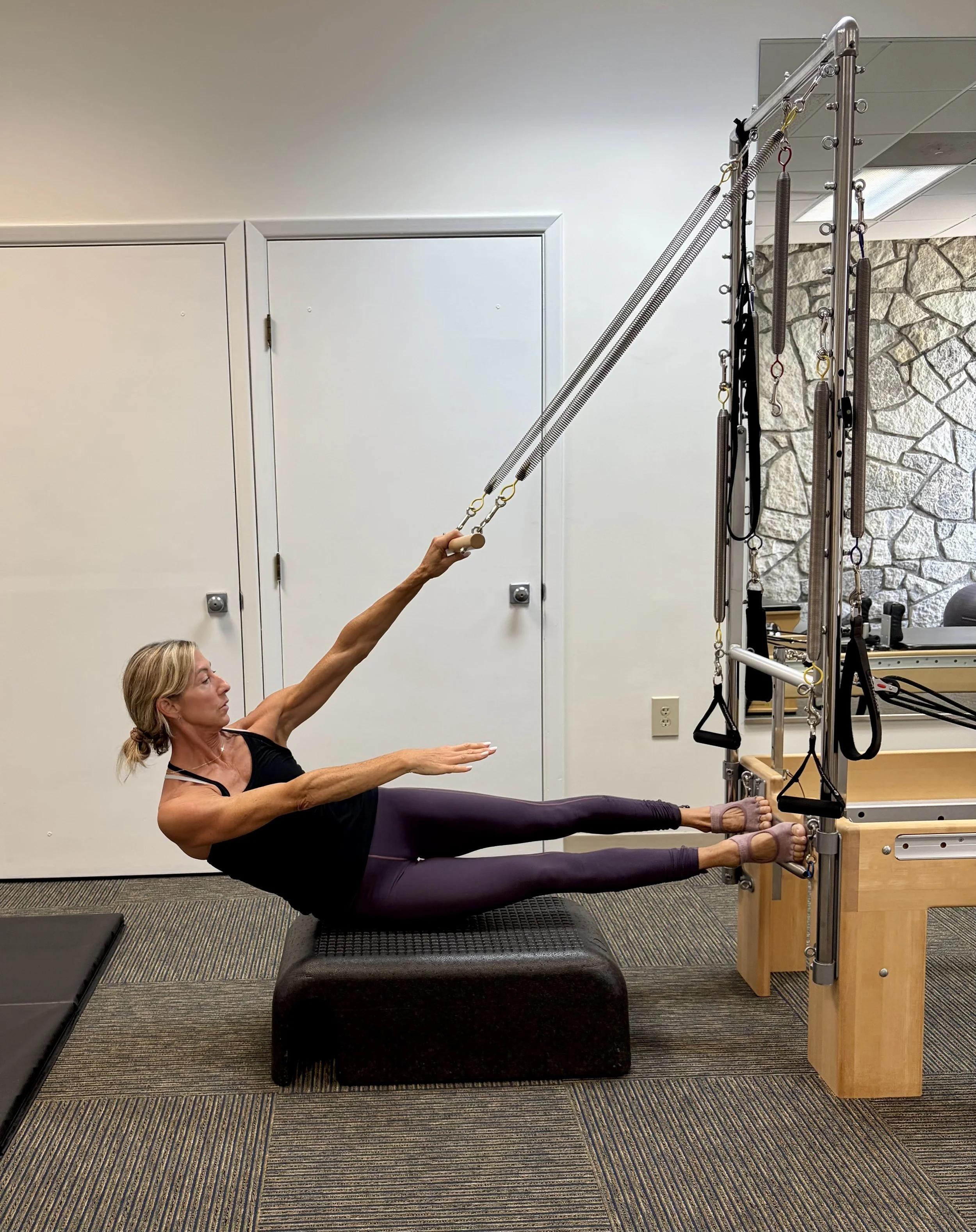 A woman in black tank top and purple leggings lying on a padded platform, performing an exercise with springs attached to a metal frame, holding onto a handle with one arm extended upward. The scene is set in a fitness or physical therapy room with a