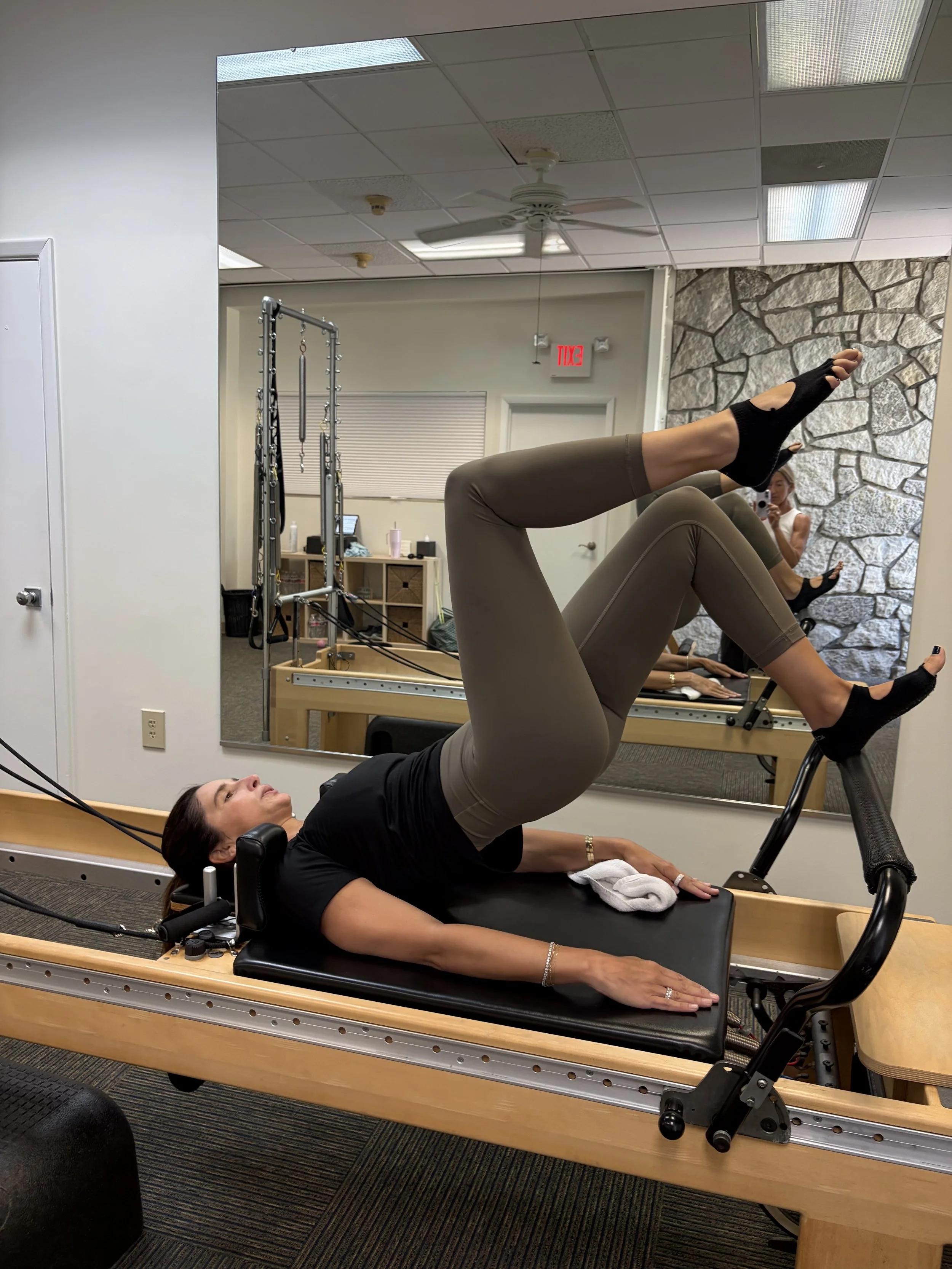 A woman performing an exercise on Pilates equipment in a studio, reflected in a mirror. She is lying on her back with her legs elevated and bent, wearing a black top, beige leggings, and black socks. The studio has a stone accent wall, a ceiling fan,