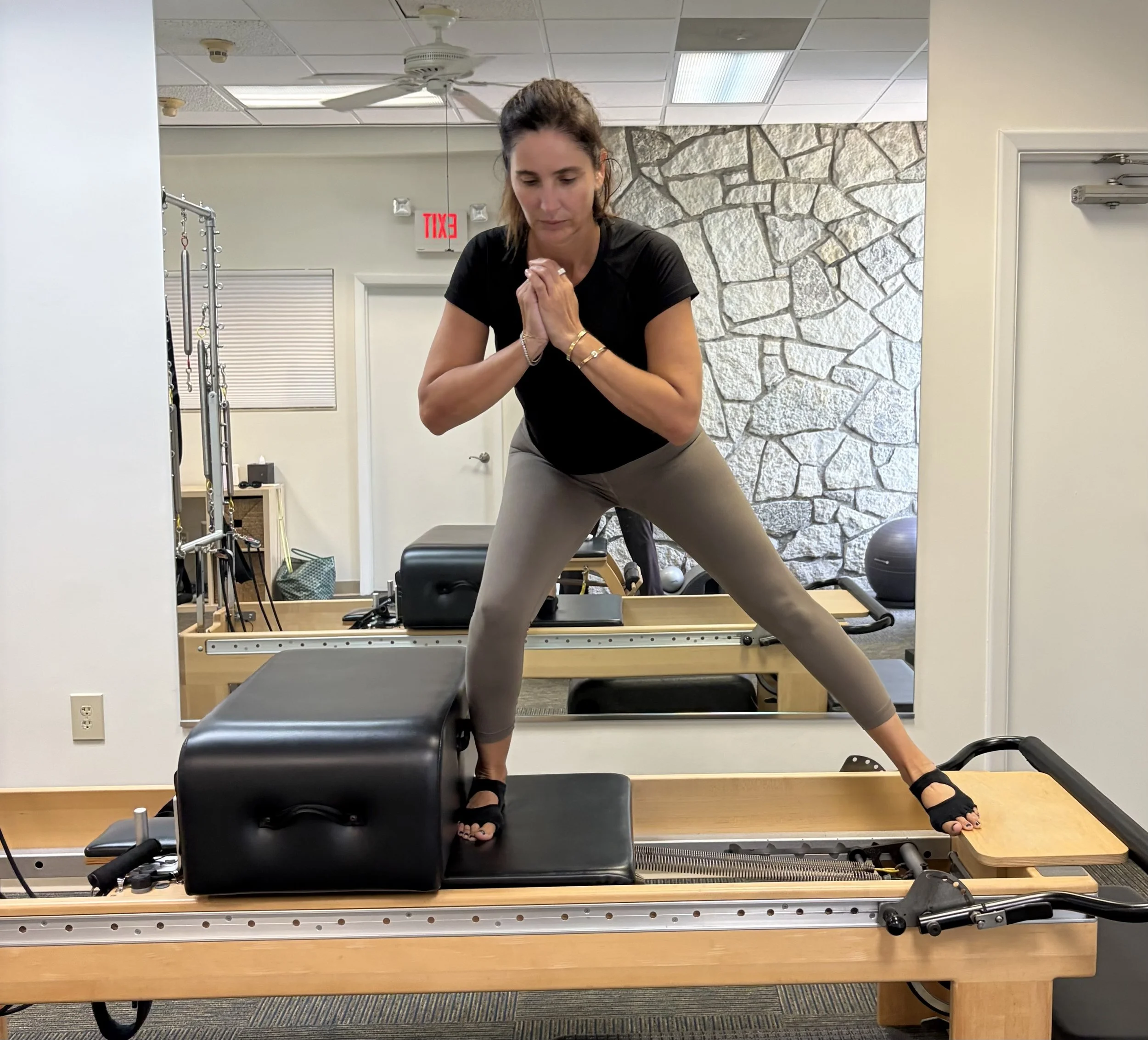 A woman practicing Pilates or yoga on a reformer machine in a fitness studio with a stone wall, a ceiling fan, and workout equipment in the background.