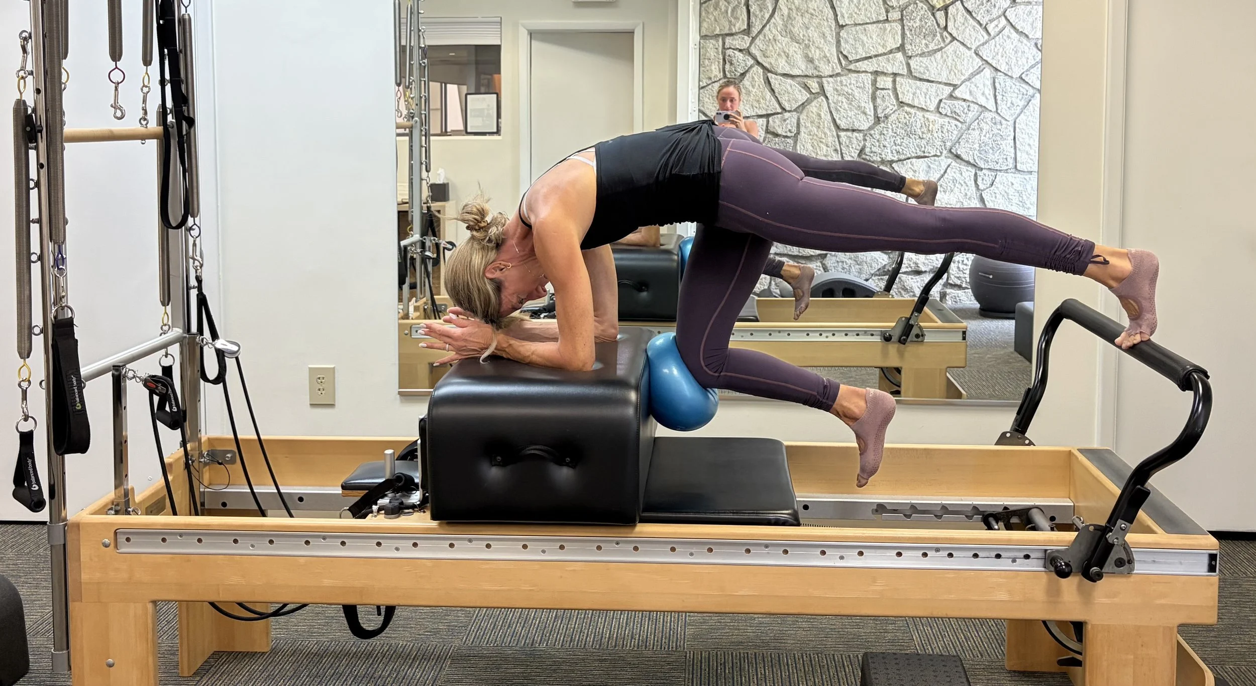 Woman practicing yoga or stretching on a Pilates reformer machine in a fitness studio with a large mirror and stone wall in the background.