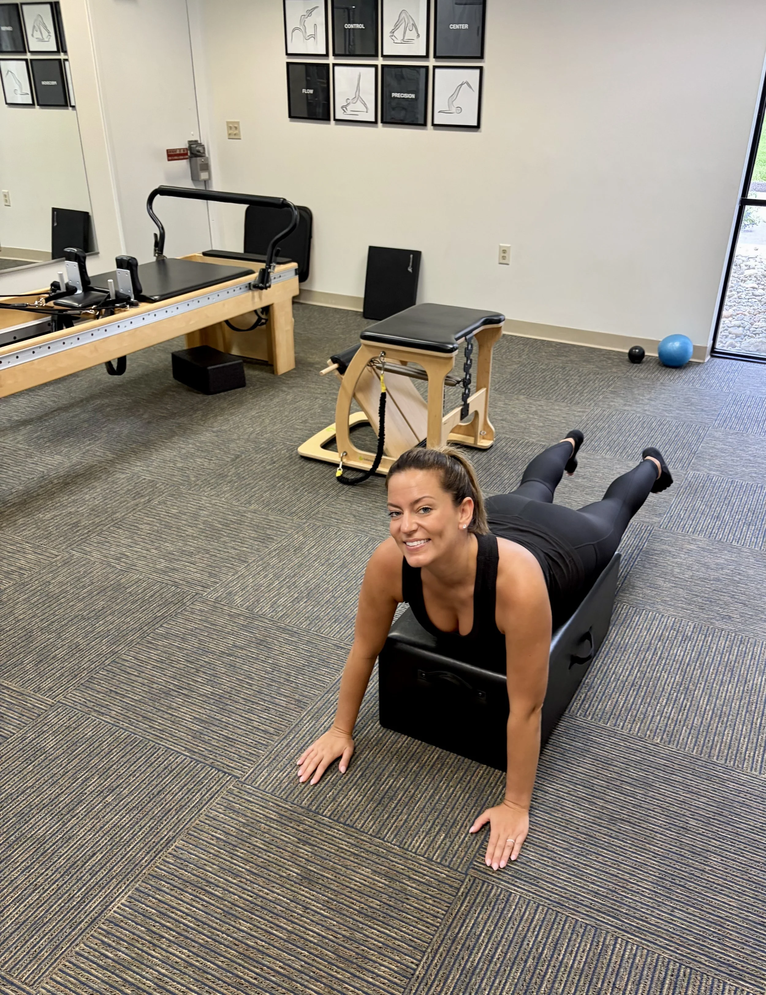 A woman doing a plank exercise on a black mat in a fitness or physical therapy room with exercise equipment, including a reformer machine and a rolling chair, visible in the background. She is smiling and looking at the camera.