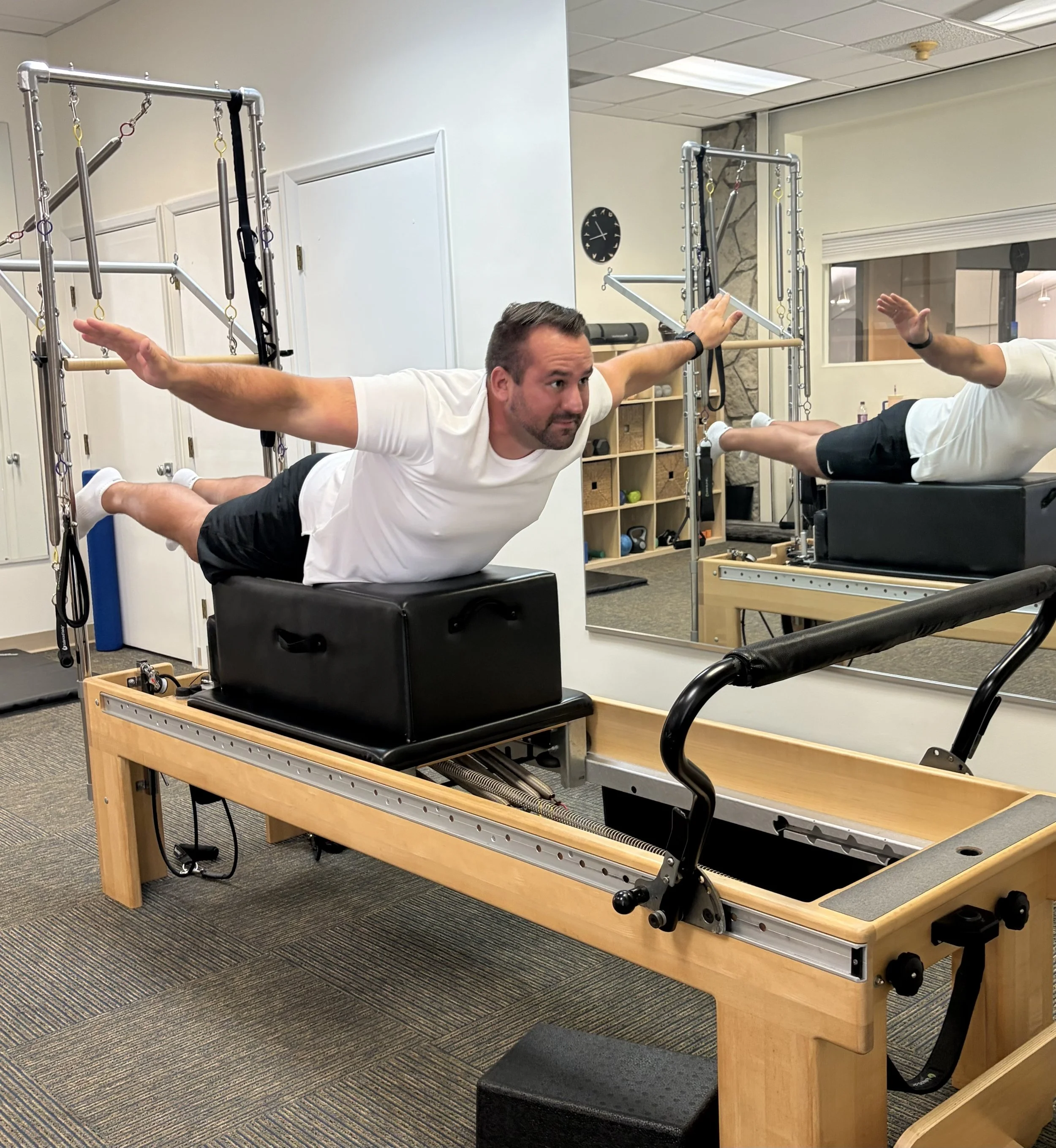 A man is performing a flying exercise on a Pilates reformer machine in a fitness studio. He is lying face down with arms and legs extended, using resistance straps.