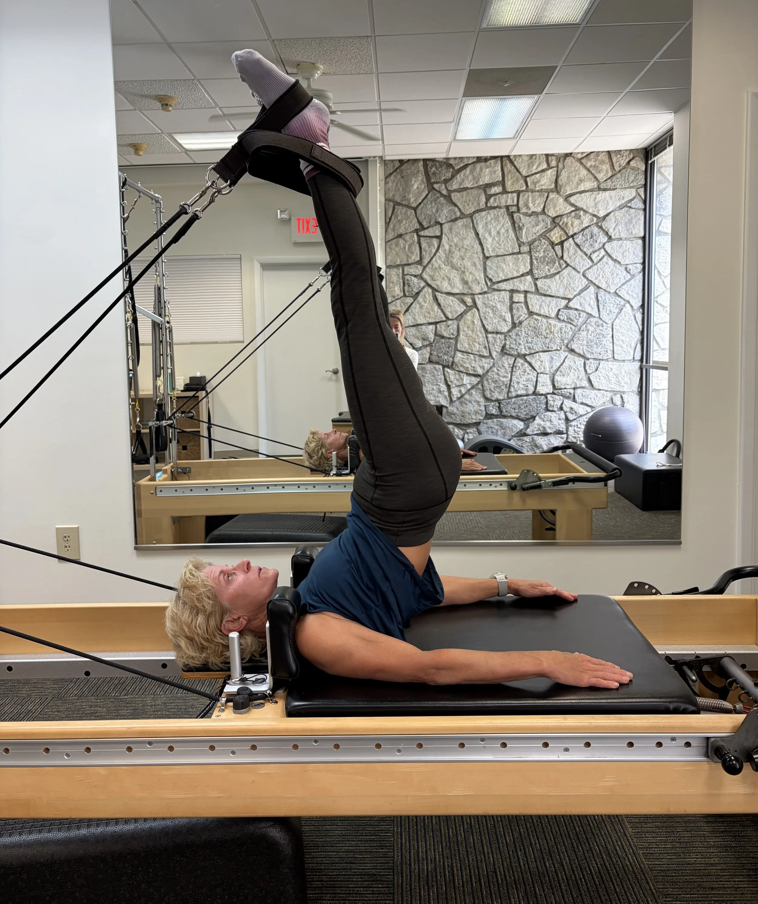 A woman lying on a Pilates reformer machine with her legs raised vertically and supported by straps in a fitness studio. The studio features a stone accent wall, large windows, and a woman in the background observing or instructing.