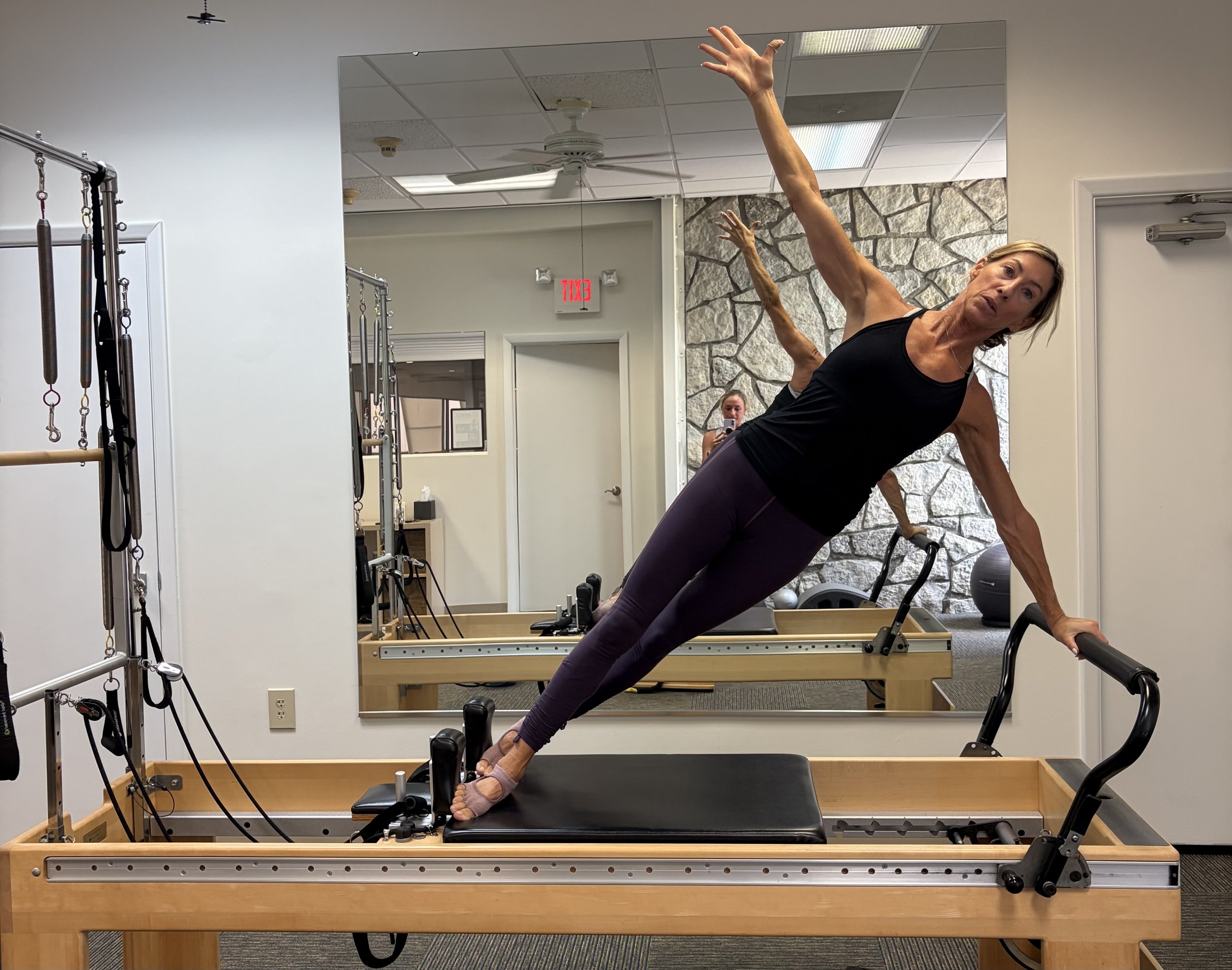A woman doing a Pilates exercise on a reformer machine in a gym, with a large mirror and stone wall in the background.