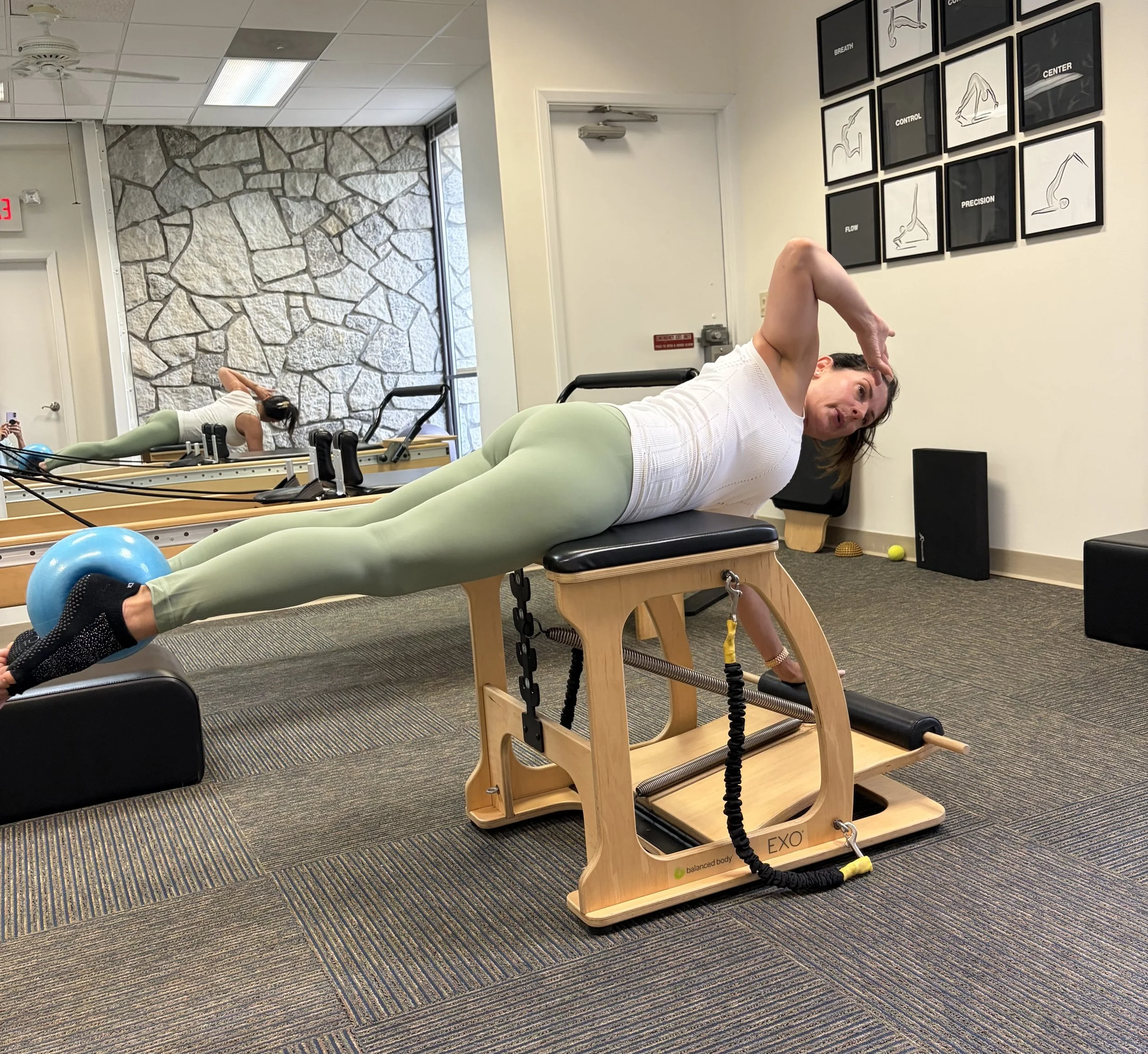 A woman doing Pilates exercises on a reformer machine in a fitness studio. She is lying on her side with one arm resting on her head, wearing a white top and light green leggings. The studio has a stone accent wall, framed fitness posters on the wall