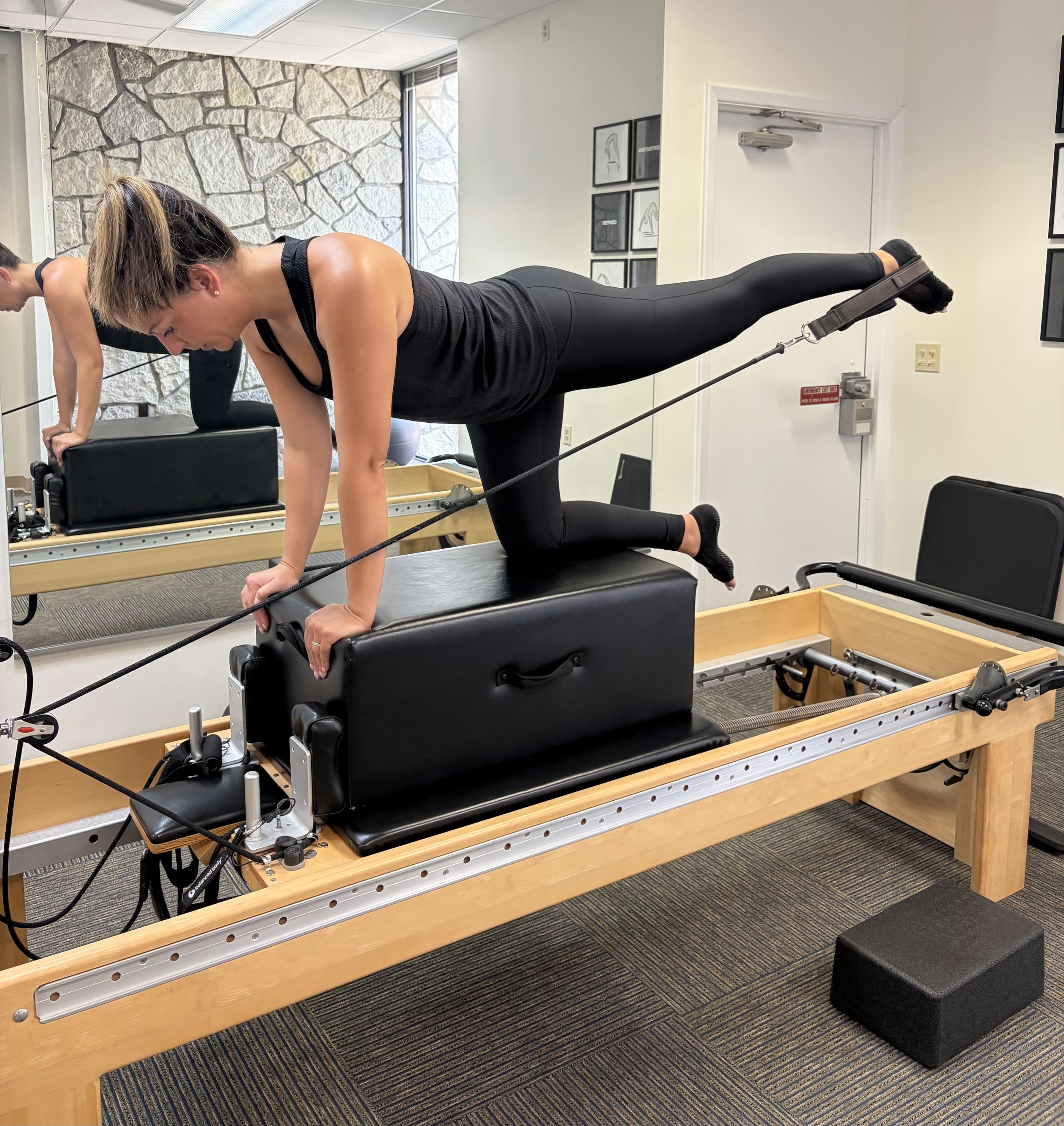 A woman in black workout clothes and a ponytail performs a Pilates exercise on a reformer machine in a fitness studio, with another person partially visible in the background.