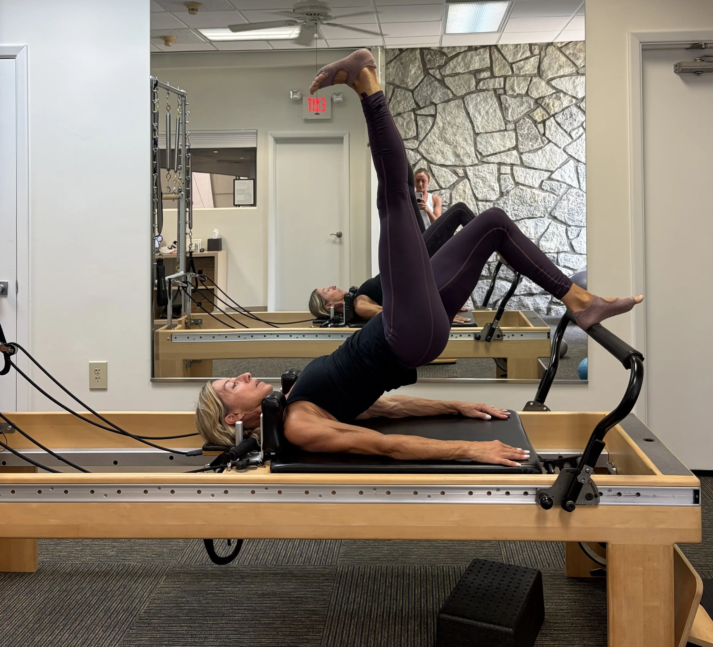 Woman lying on a Pilates reformer machine with her legs raised vertical, exercising, in a room with a stone accent wall and a large mirror reflecting her and the room.
