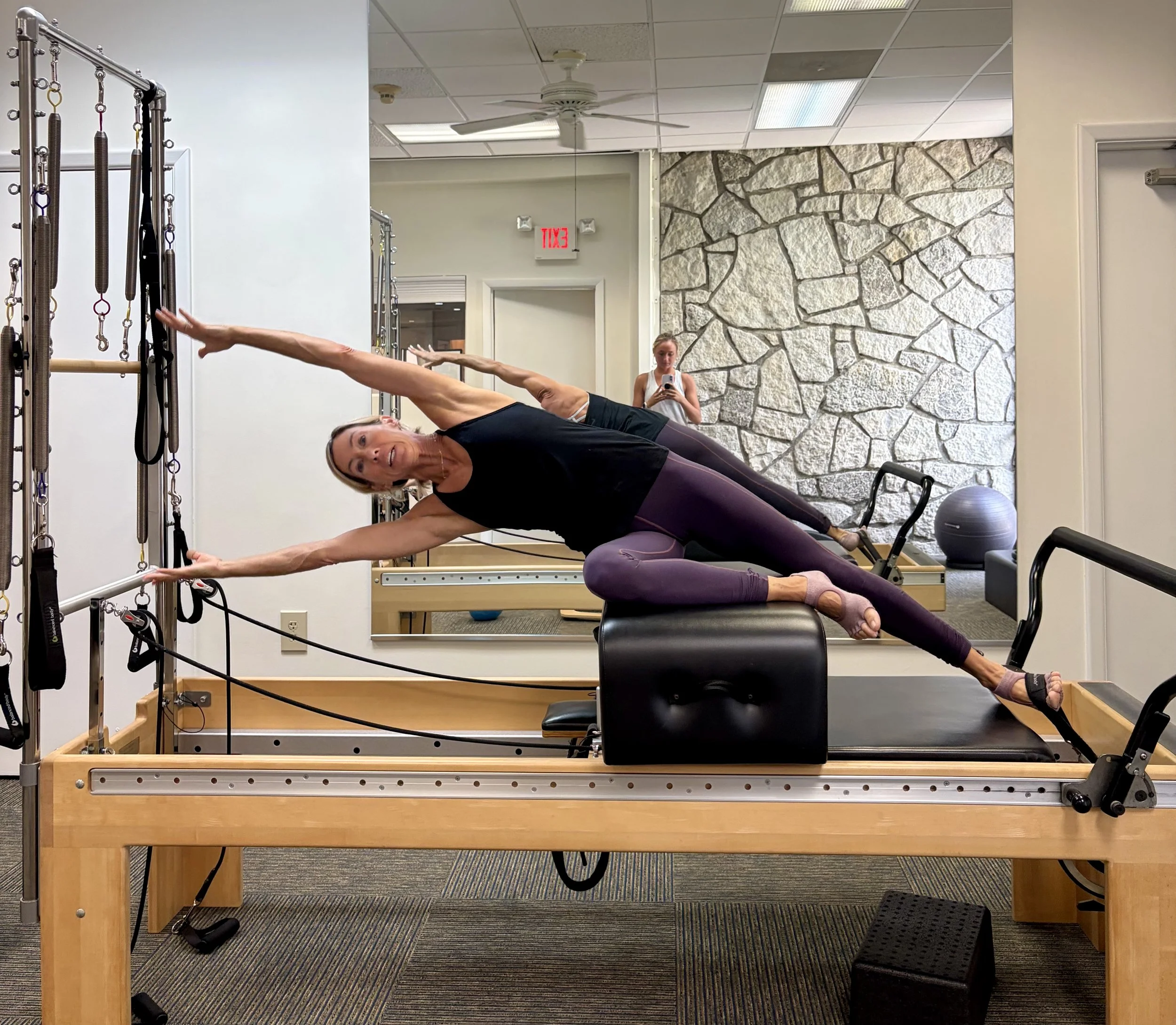 Woman performing a side plank exercise on a Pilates reformer machine in a fitness studio, with a woman taking a photo in the background.