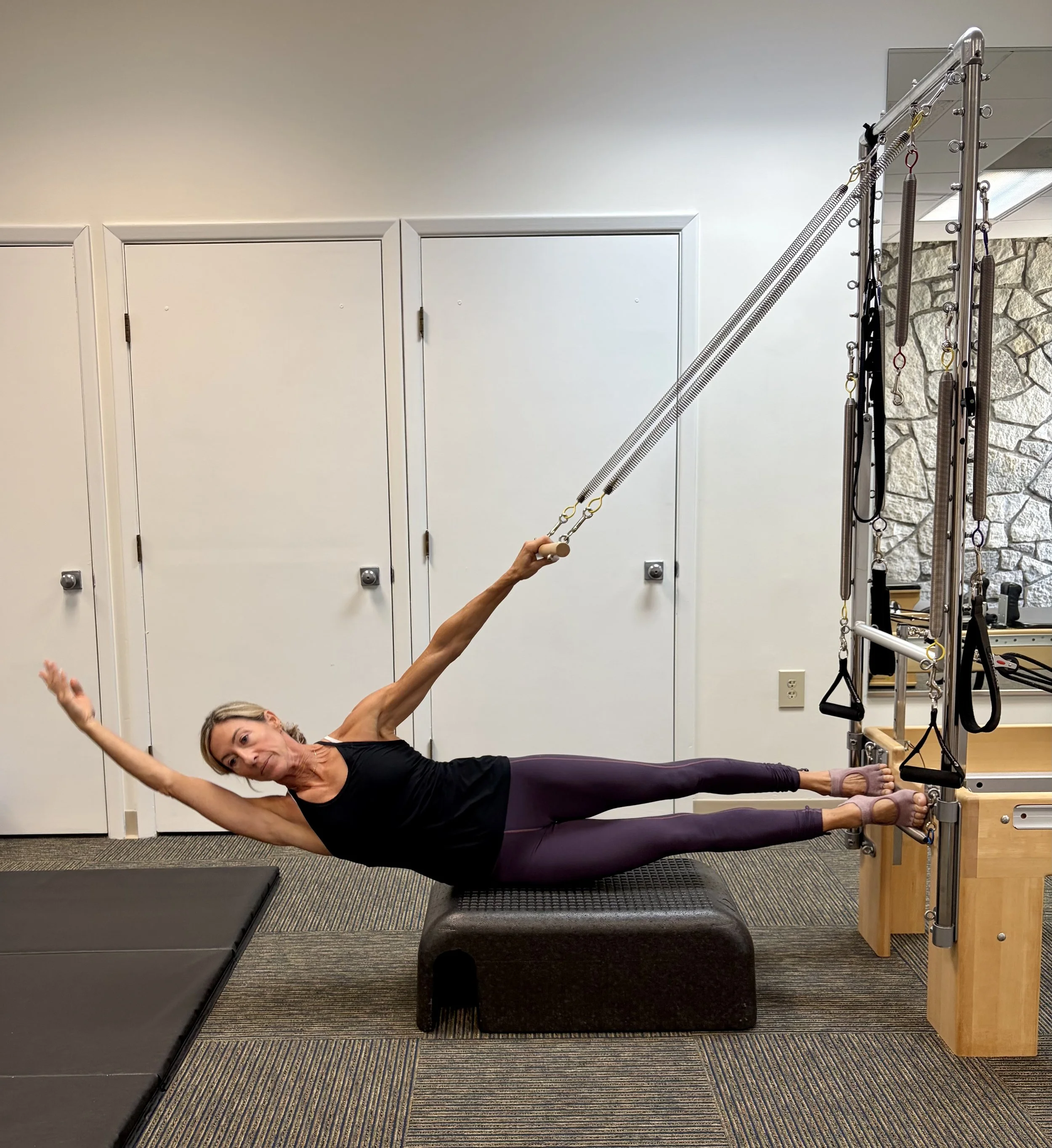 A woman doing a side plank exercise on a padded mat in a fitness studio. She is holding a dental floss-like exercise tool, stabilizing her body.