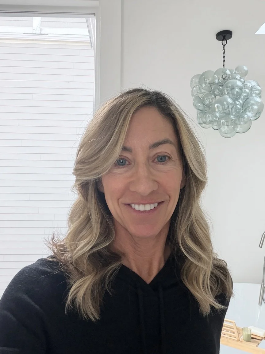 A woman with blonde wavy hair taking a selfie in a modern, brightly lit room with a white wall, window, and decorative hanging chandelier.