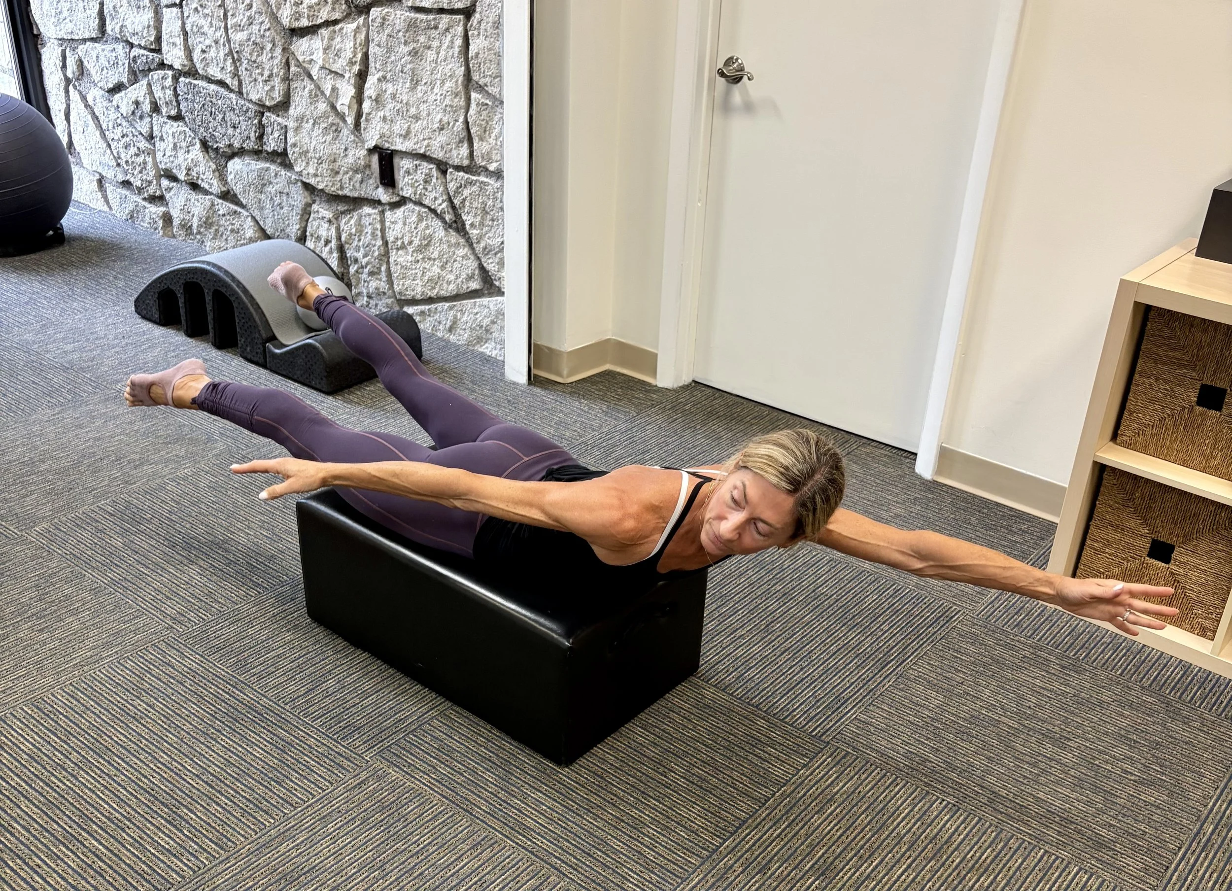 A woman is lying on a black exercise bench, stretching with her arm extended forward and legs in a raised position, performing a workout in a room with a stone wall, closed door, beige walls, and a wooden shelving unit.