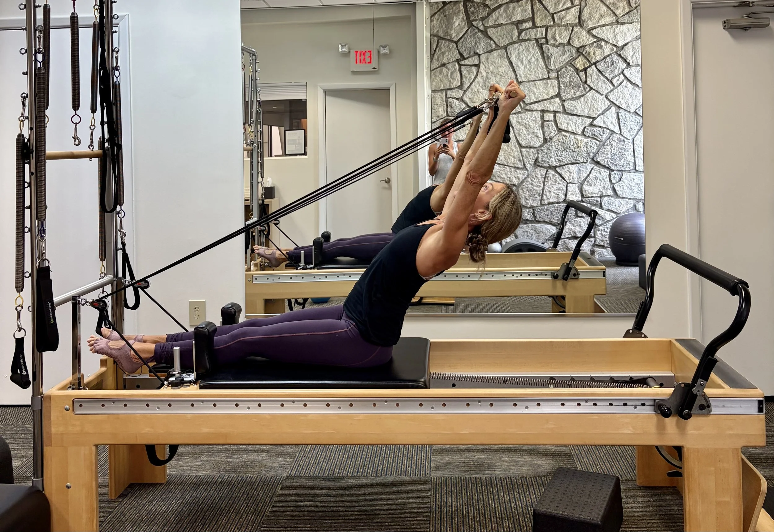 Two women exercising on Pilates reformer machines in a fitness studio, with a stone accent wall and a mirror reflecting a person taking their photo.