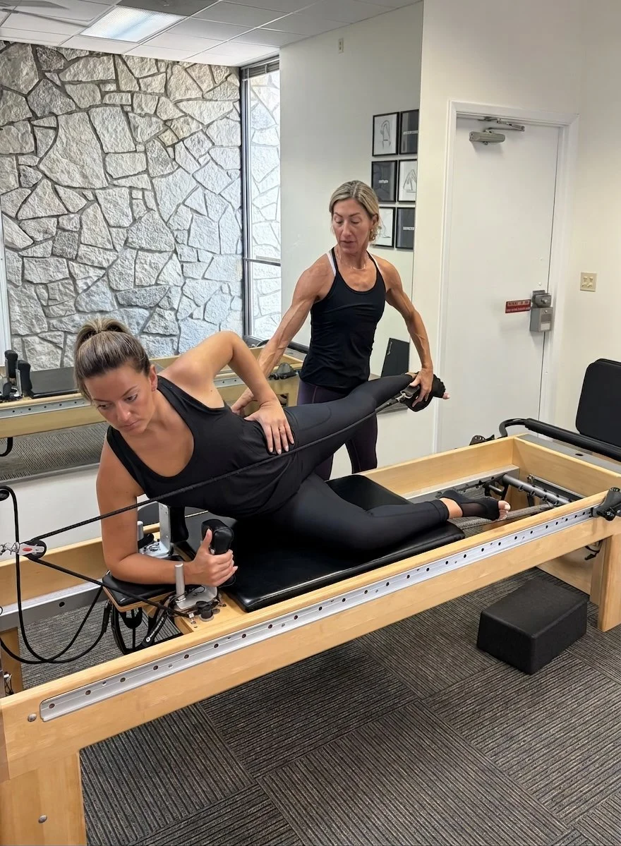 A woman is lying on an advanced Pilates reformer machine, exercising with a resistance band while a female instructor assists her. The instructor stands beside her, guiding her movement in a fitness studio with a stone accent wall and framed artwork.