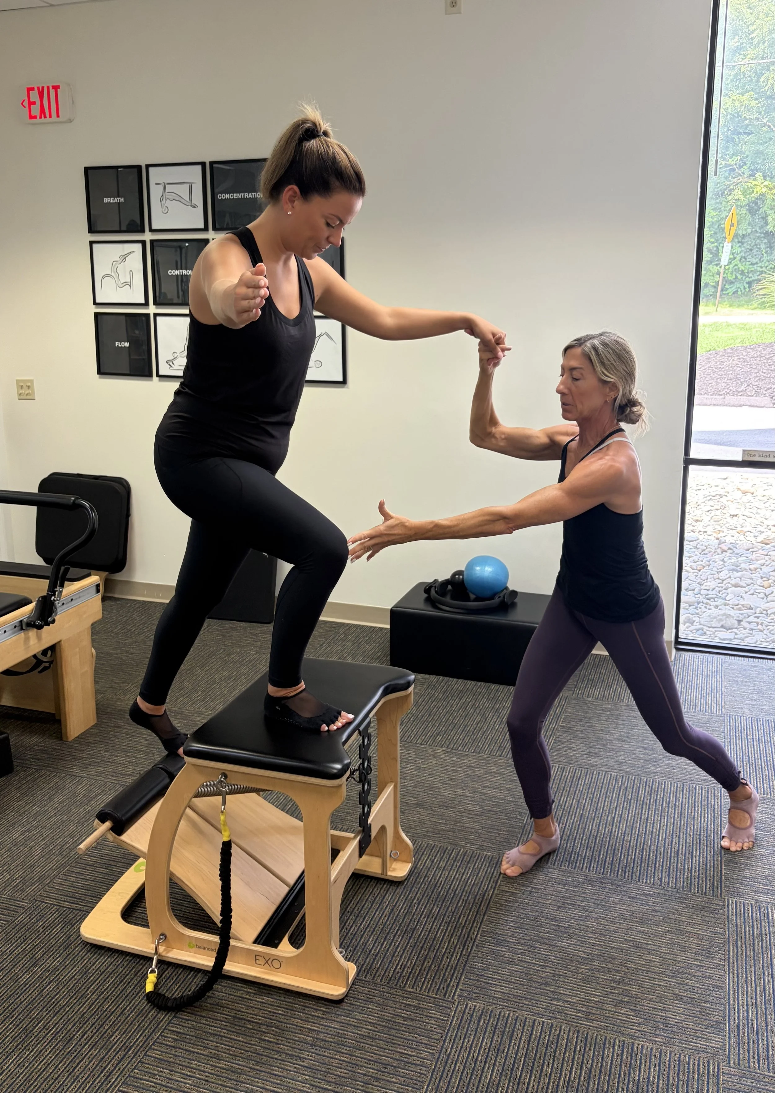 Two women are exercising together in a room with large windows. One is on a Pilates reformer machine, balancing on her hands and feet with resistance bands, while the other assists her by holding her arm. The room has framed posters on the wall and e