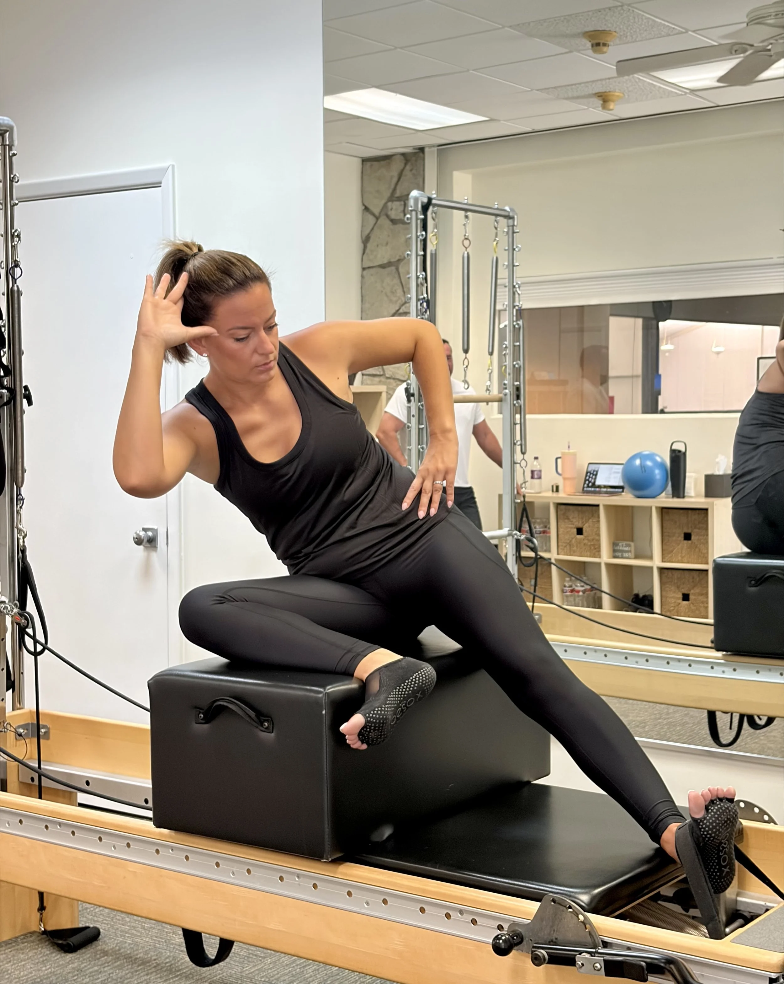 Woman doing physical therapy exercises on a specialized chair in a therapy room with exercise equipment and a large mirror.