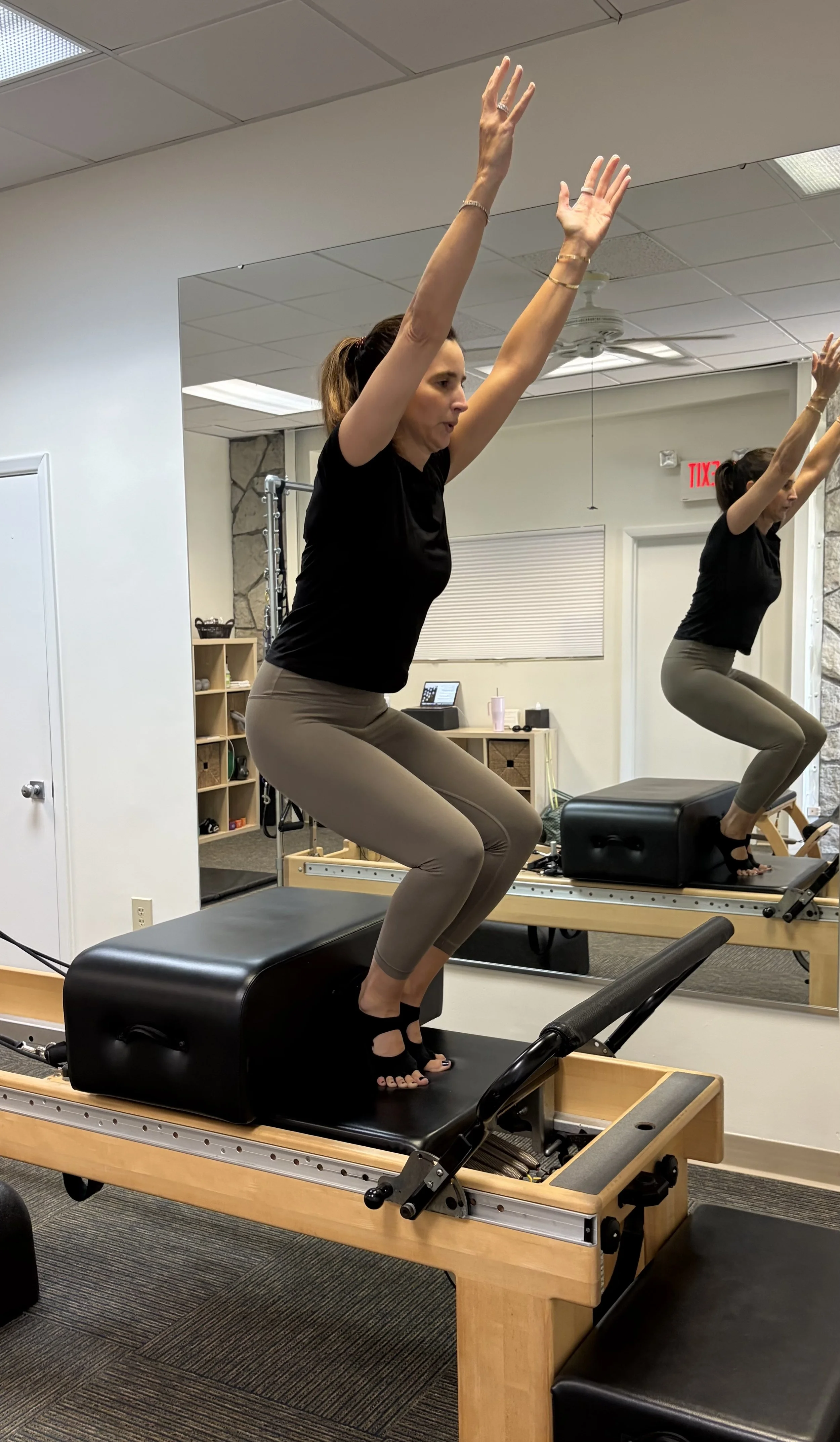 Woman exercising on Pilates reformer machine in a fitness studio, reflected in a large wall mirror.