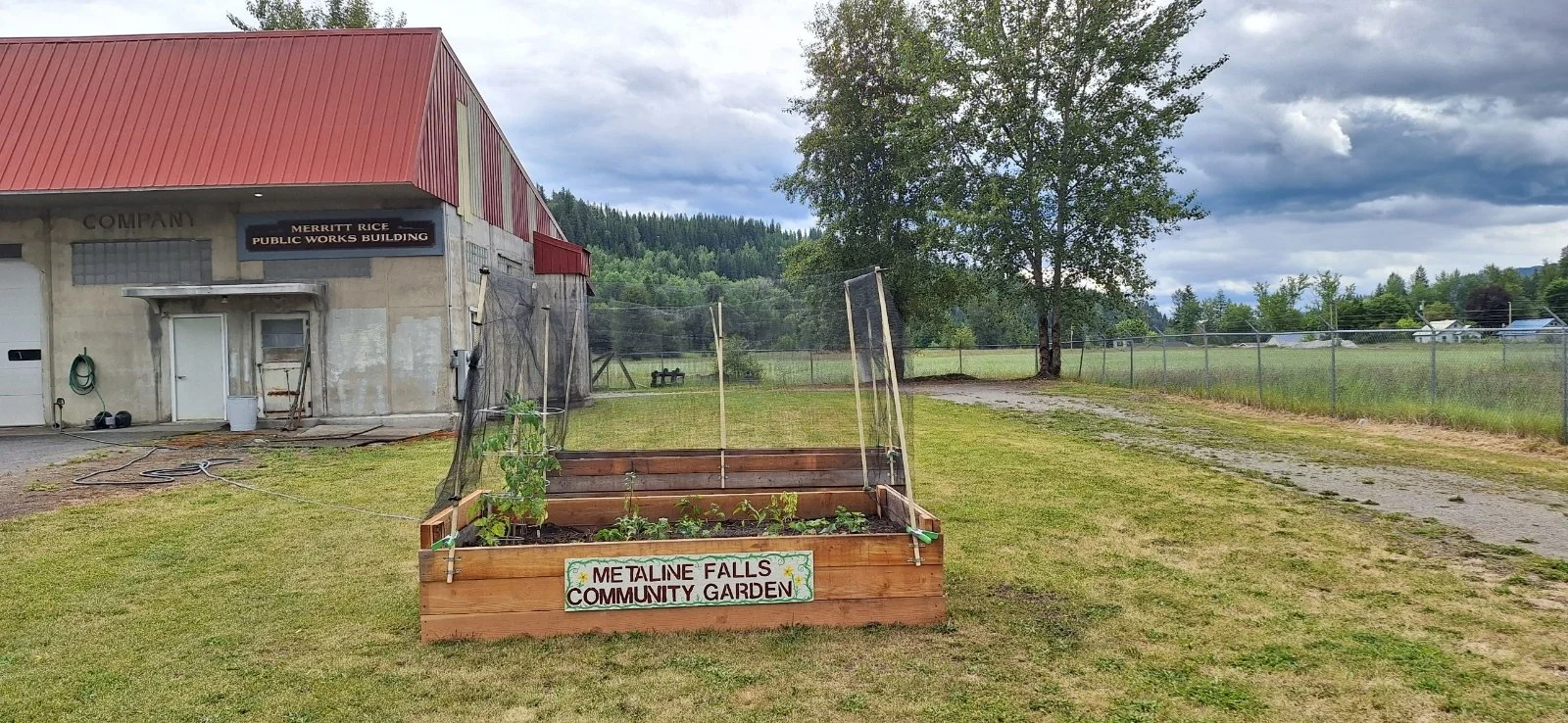 Raised garden beds with a hand-painted sign that says "Metaline Falls Community Garden." Behind and to the right of the beds is a Merritt Rice Public Works Building. in the distance is a green forest.