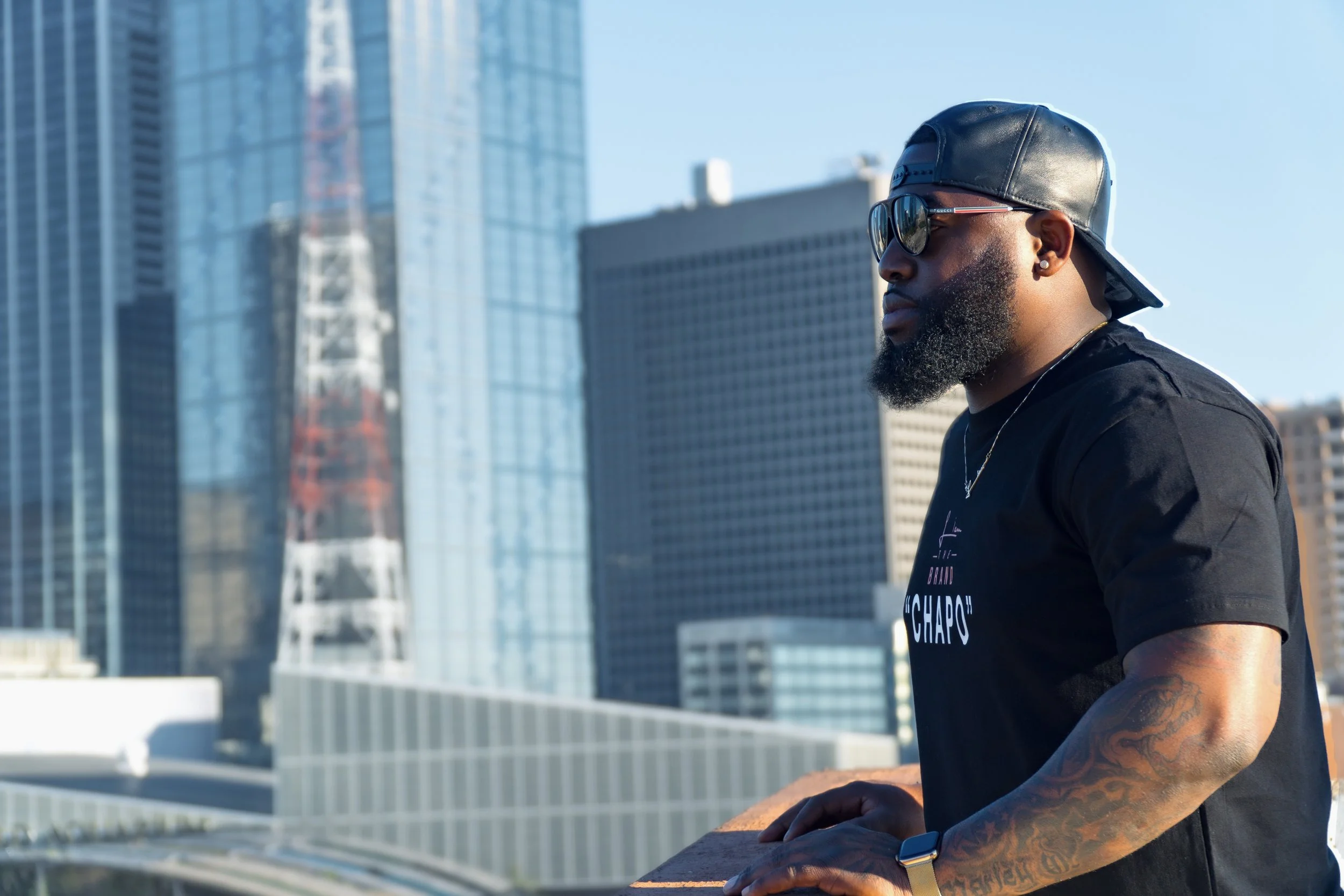 A man with a beard wearing sunglasses, a black leather cap backwards, and a black T-shirt, standing on an urban rooftop with skyscrapers in the background.