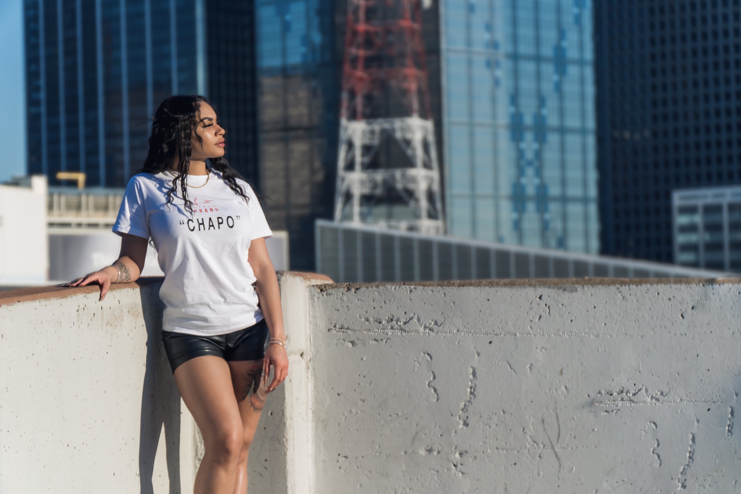 A woman with long dark hair standing on a rooftop with a cityscape background, wearing a white t-shirt with 'CHAPO' written on it and black shorts, looking to the side.