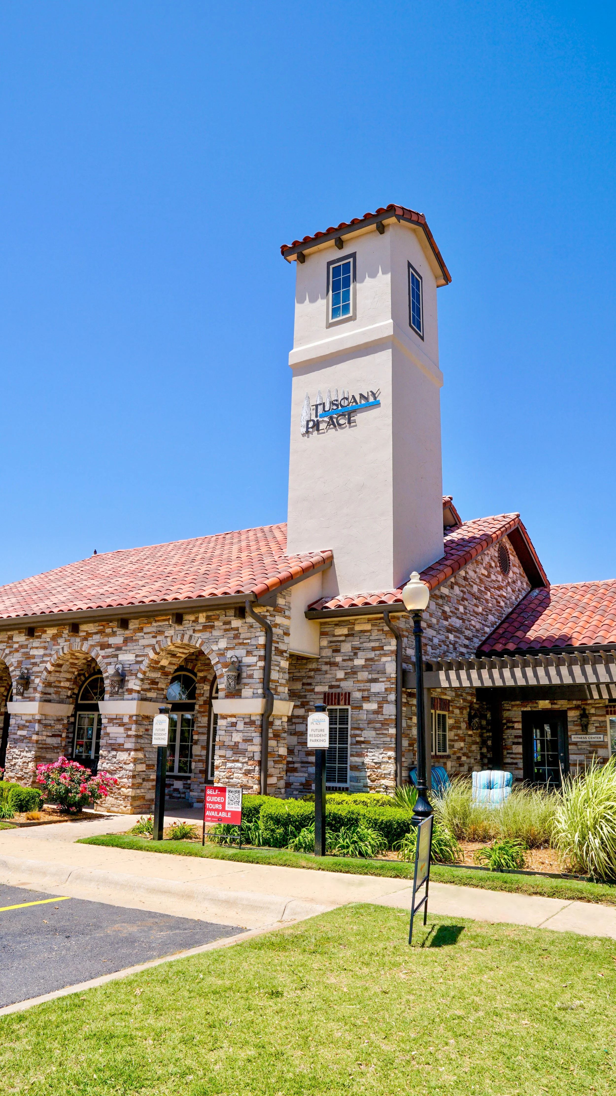 A building with a stone facade and a red tiled roof, featuring a white tower with a sign that reads 'Tuscany Place' crossed out. There are signs indicating parking for residents and a red sign offering self-guided tours. The area is landscaped with g