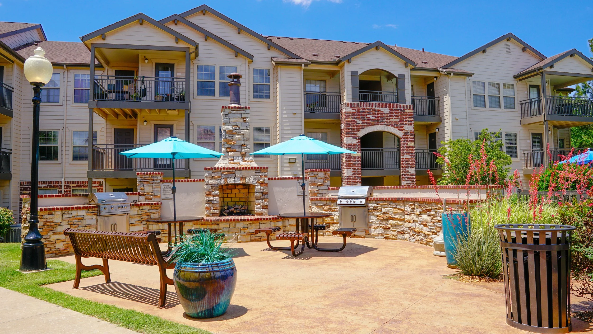 Outdoor communal area with a stone fireplace, two blue umbrellas, BBQ grills, a bench, planters, and a multi-story apartment building with balconies in the background.
