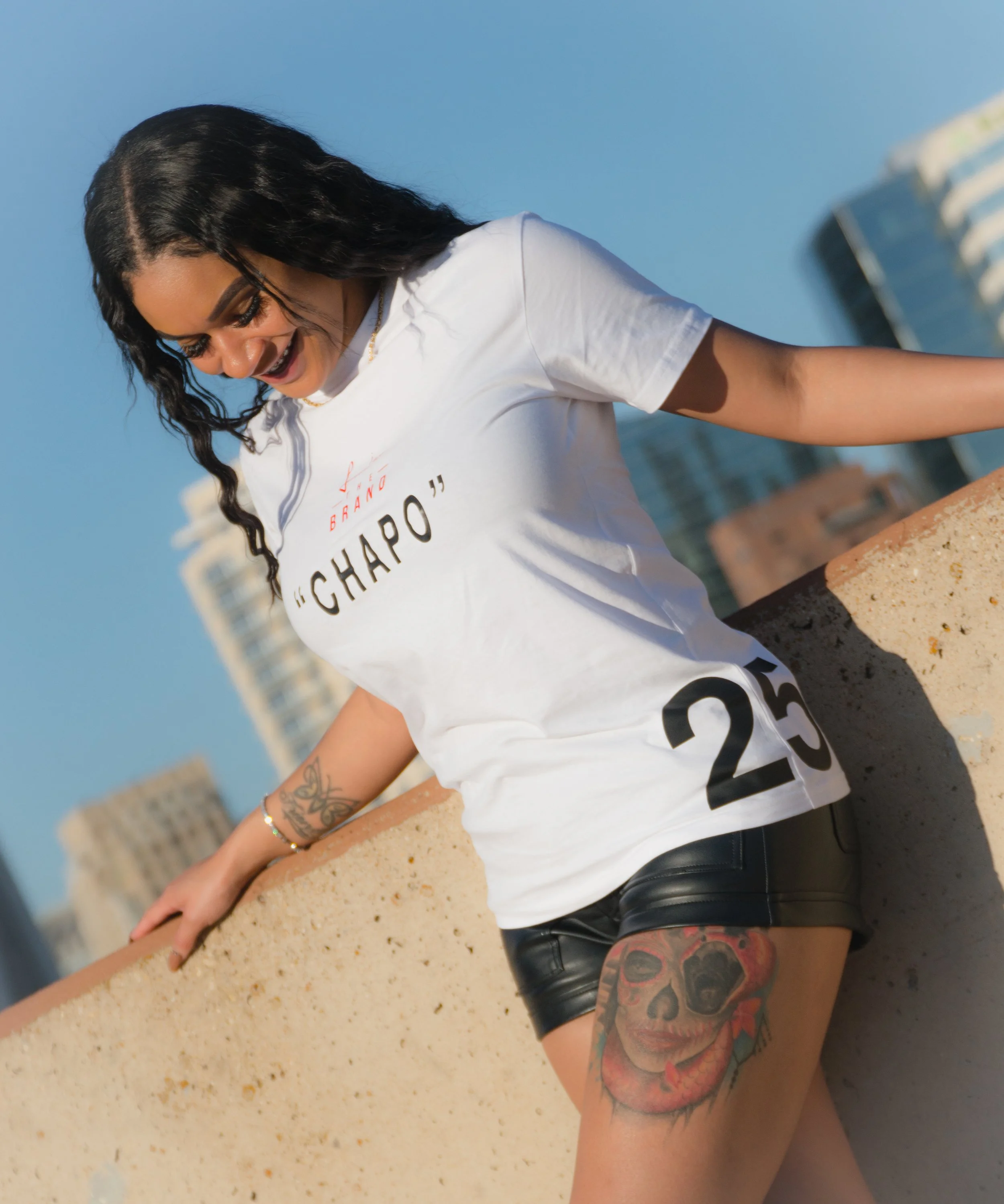 Young woman with black wavy hair wearing a white T-shirt and black leather shorts, leaning on a ledge outdoors with city buildings in the background, smiling.