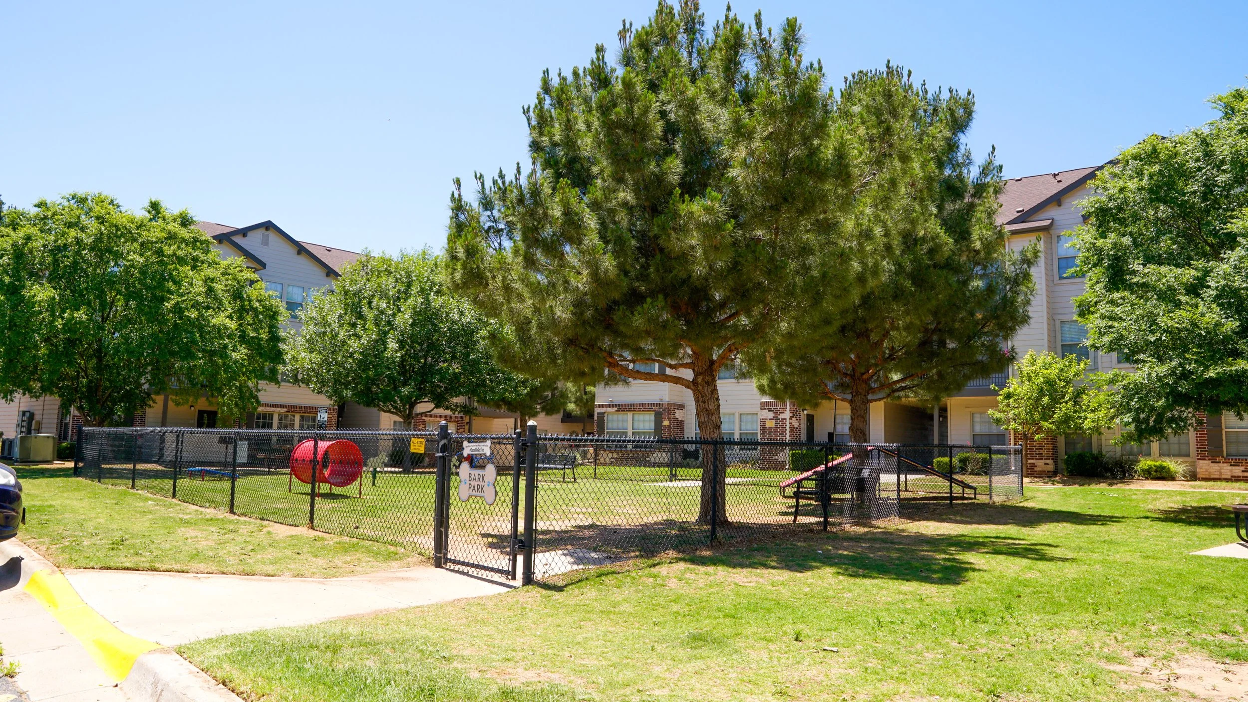 A fenced dog park with trees, a playground slide, and a sign that says 'Bark Park' in a residential apartment complex.