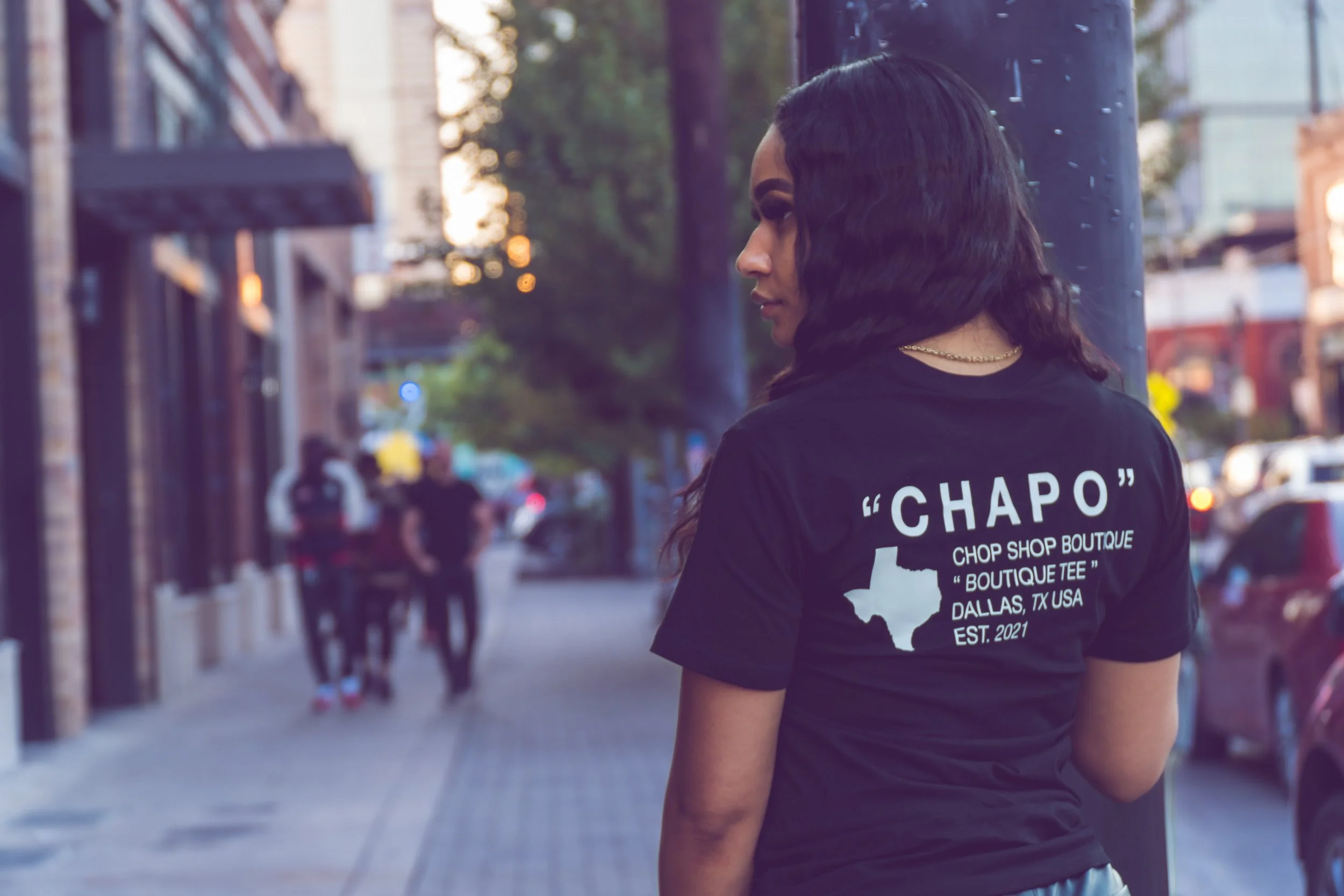 A woman with dark hair standing on a city sidewalk near a lamppost, wearing a black T-shirt with white text and a map of Texas, with people walking in the background.