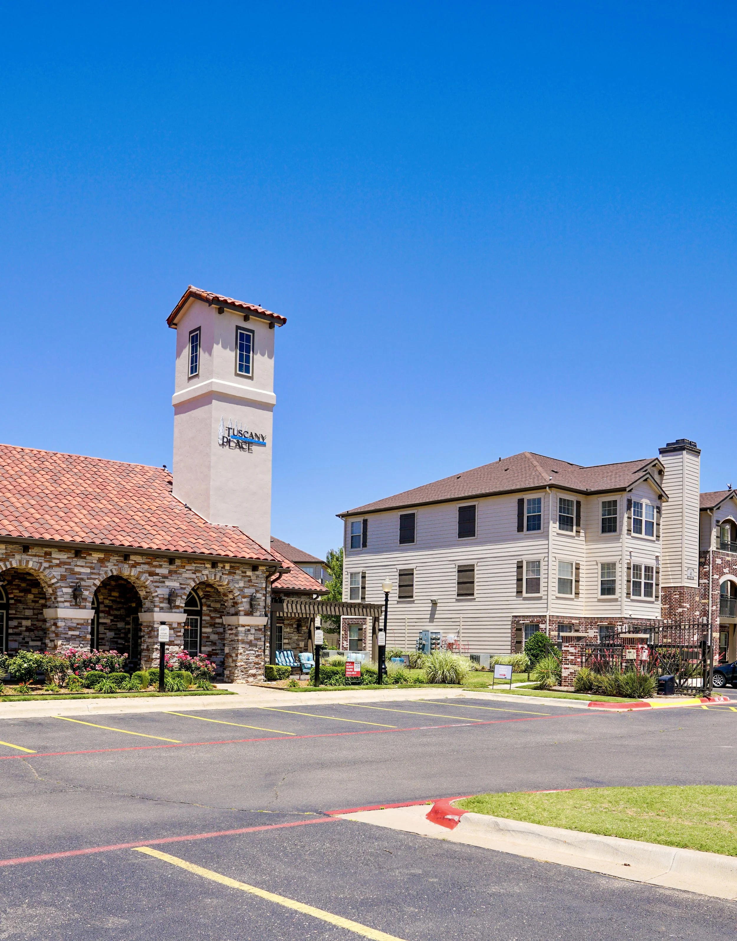 A parking lot with a building labeled Toscana Place and a tall tower with a red roof against a bright blue sky.