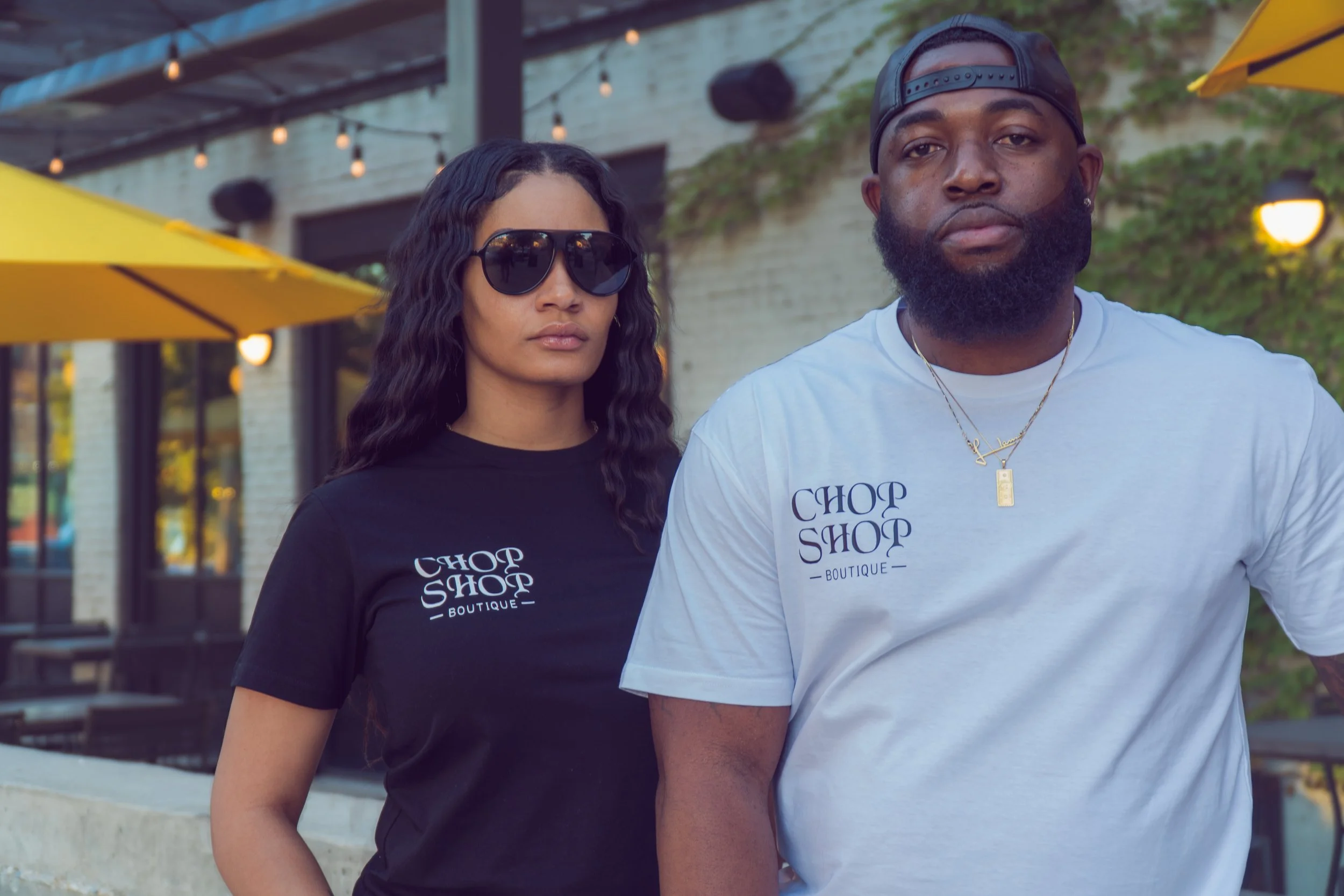 A woman and a man stand outdoors, wearing matching 'Chop Shop Boutique' T-shirts. The woman is wearing black sunglasses and has long, wavy hair. The man is wearing a backward cap, has a beard, and gold jewelry. There's a yellow umbrella and a brick b